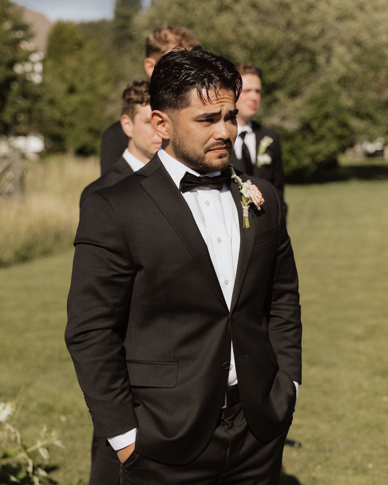A groom in a classic black tuxedo and bow tie stands outdoors on a sunny day during a wedding ceremony.