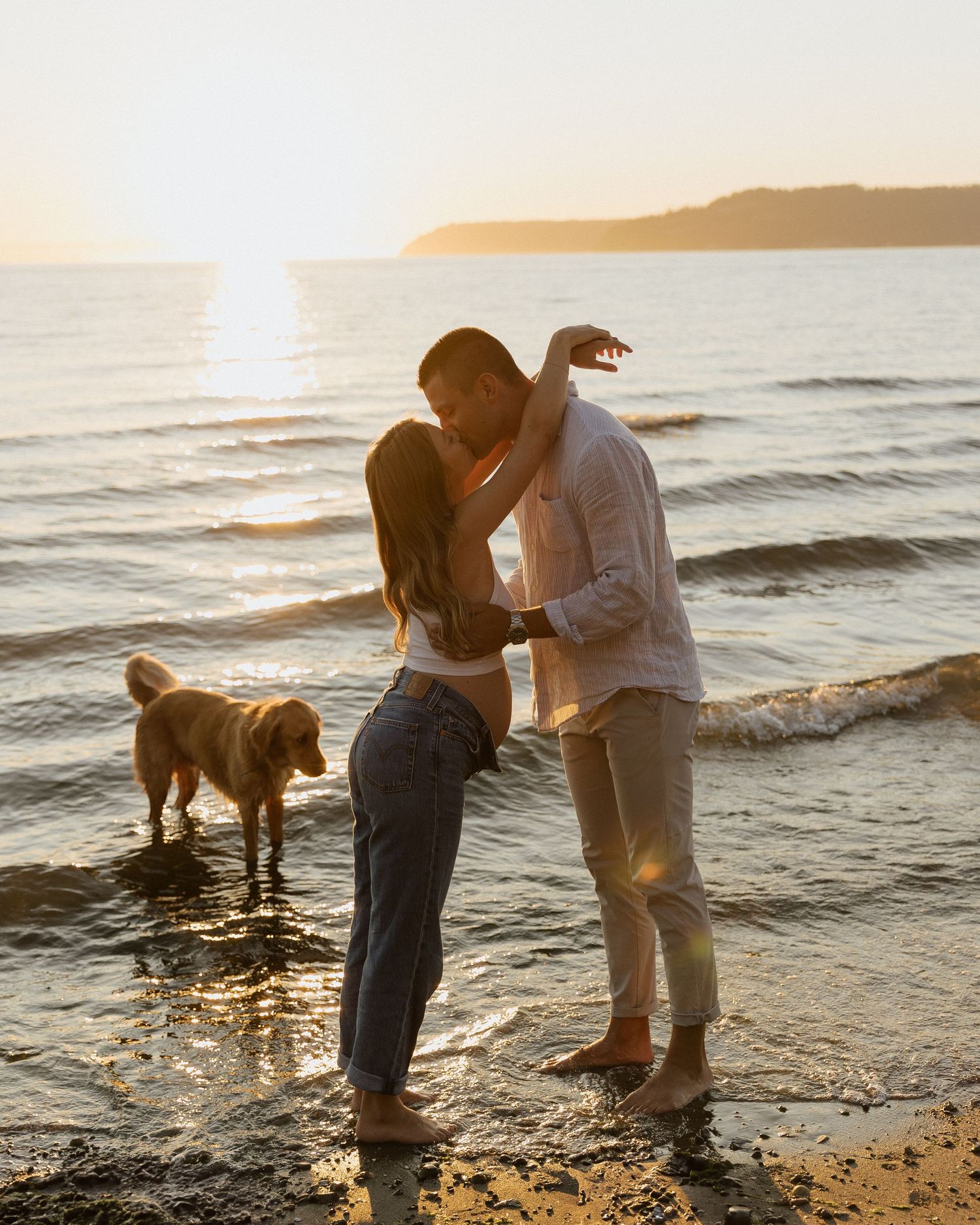 Romantic couple shares tender moment by the water with dog nearby during sunset.