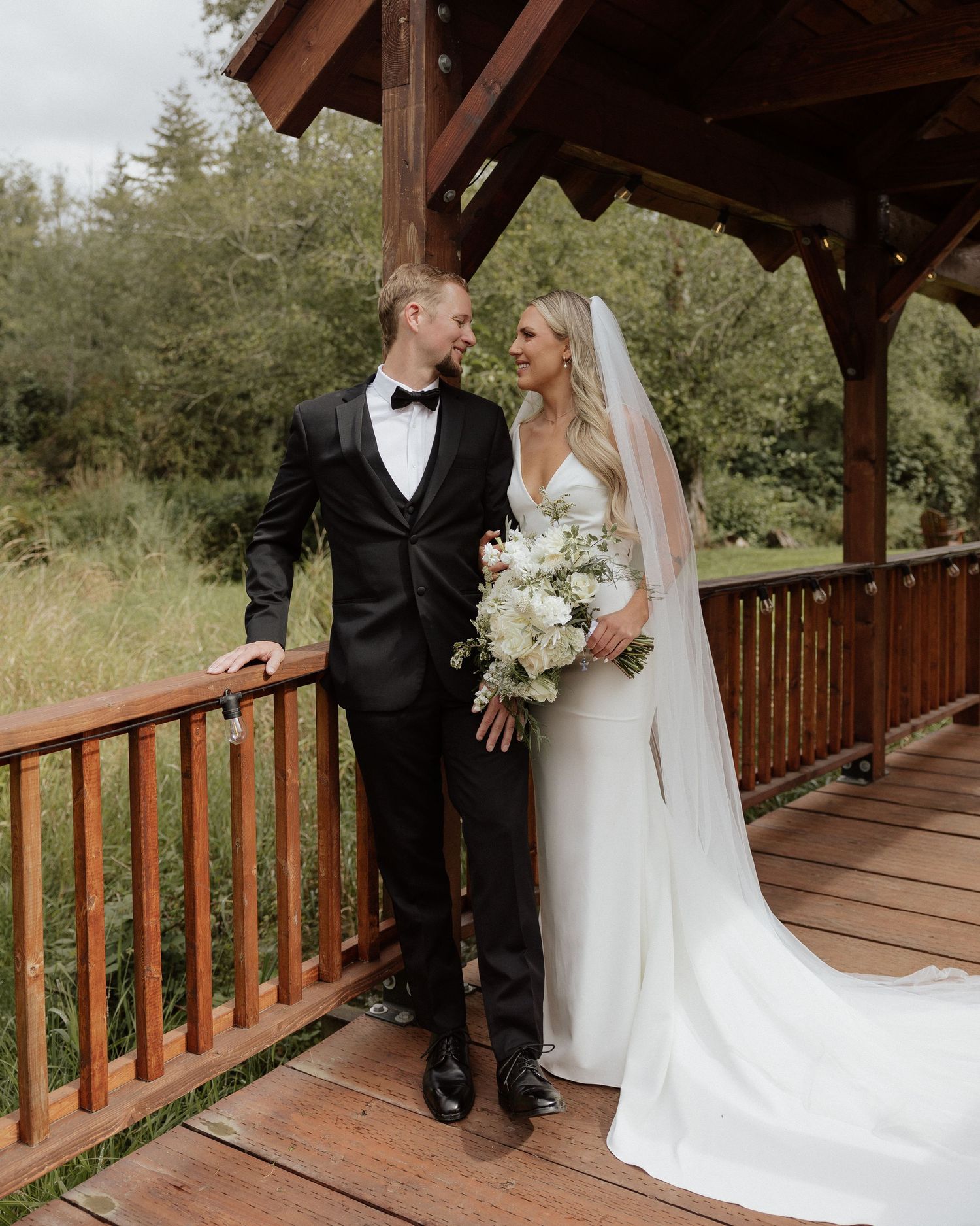 Elegant wedding couple posing on covered wooden bridge with white floral bouquet.