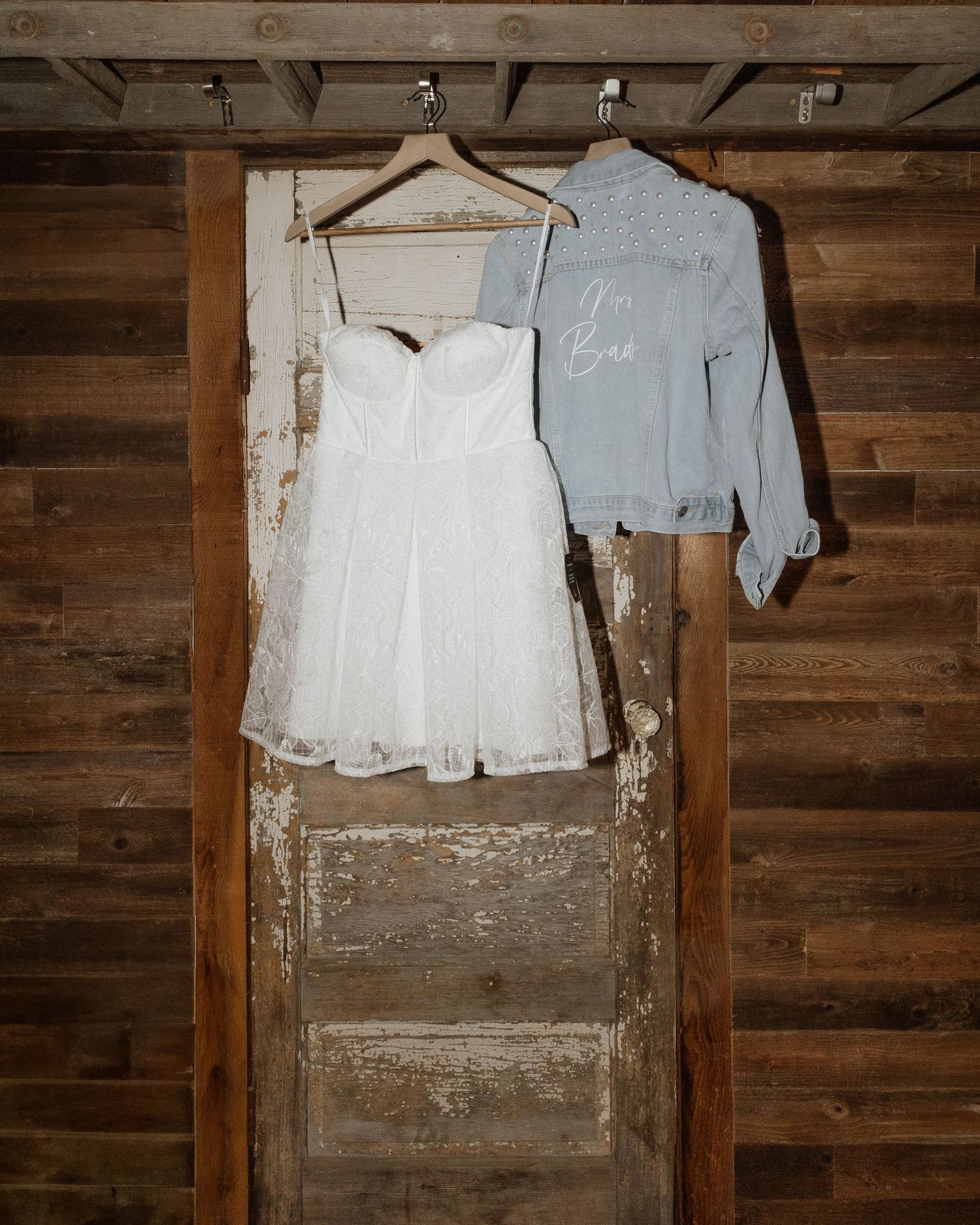 A white wedding dress and light blue jacket hanging on wooden hangers against a rustic wooden wall.