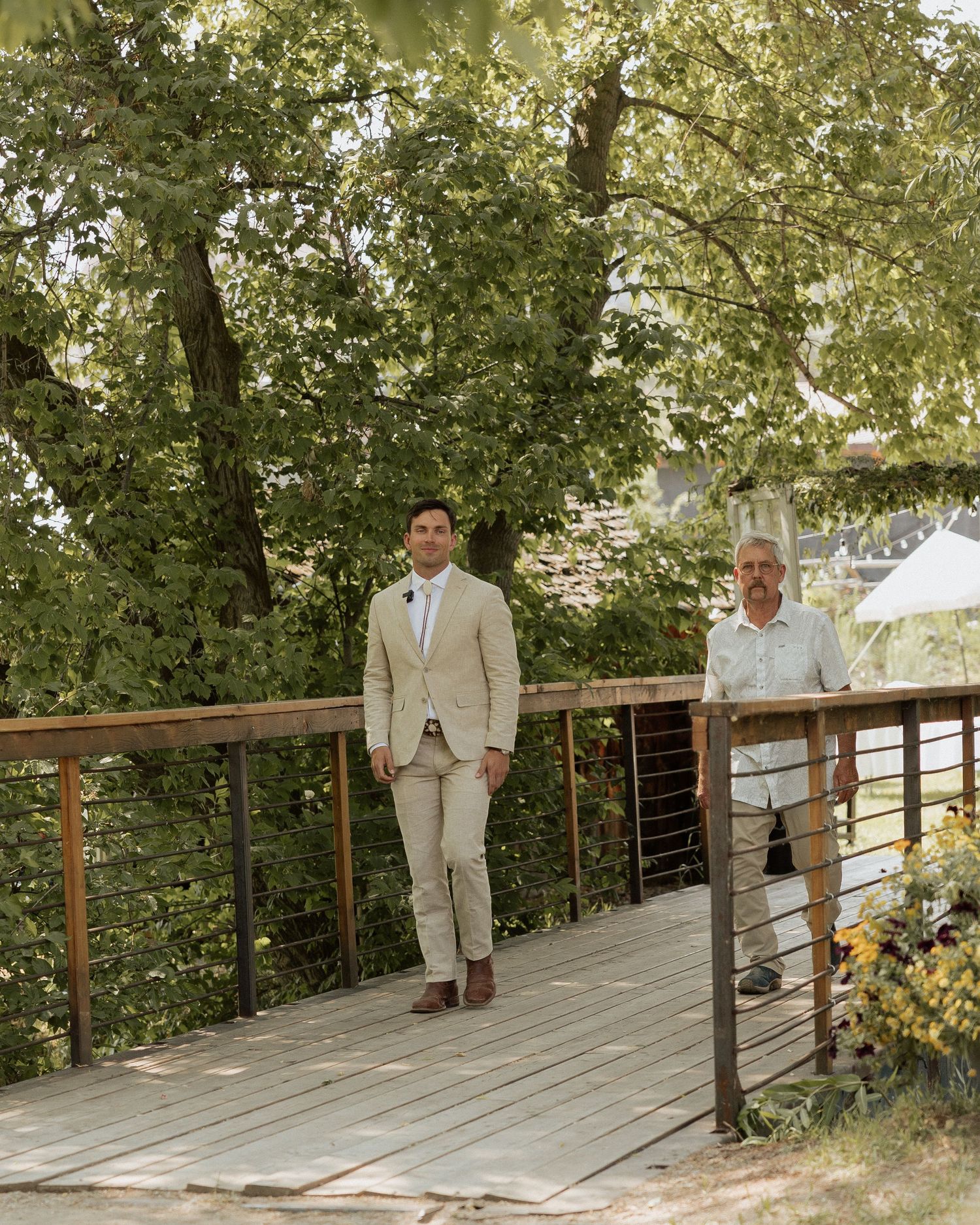Two people in light colored suits stand on wooden deck surrounded by lush greenery.