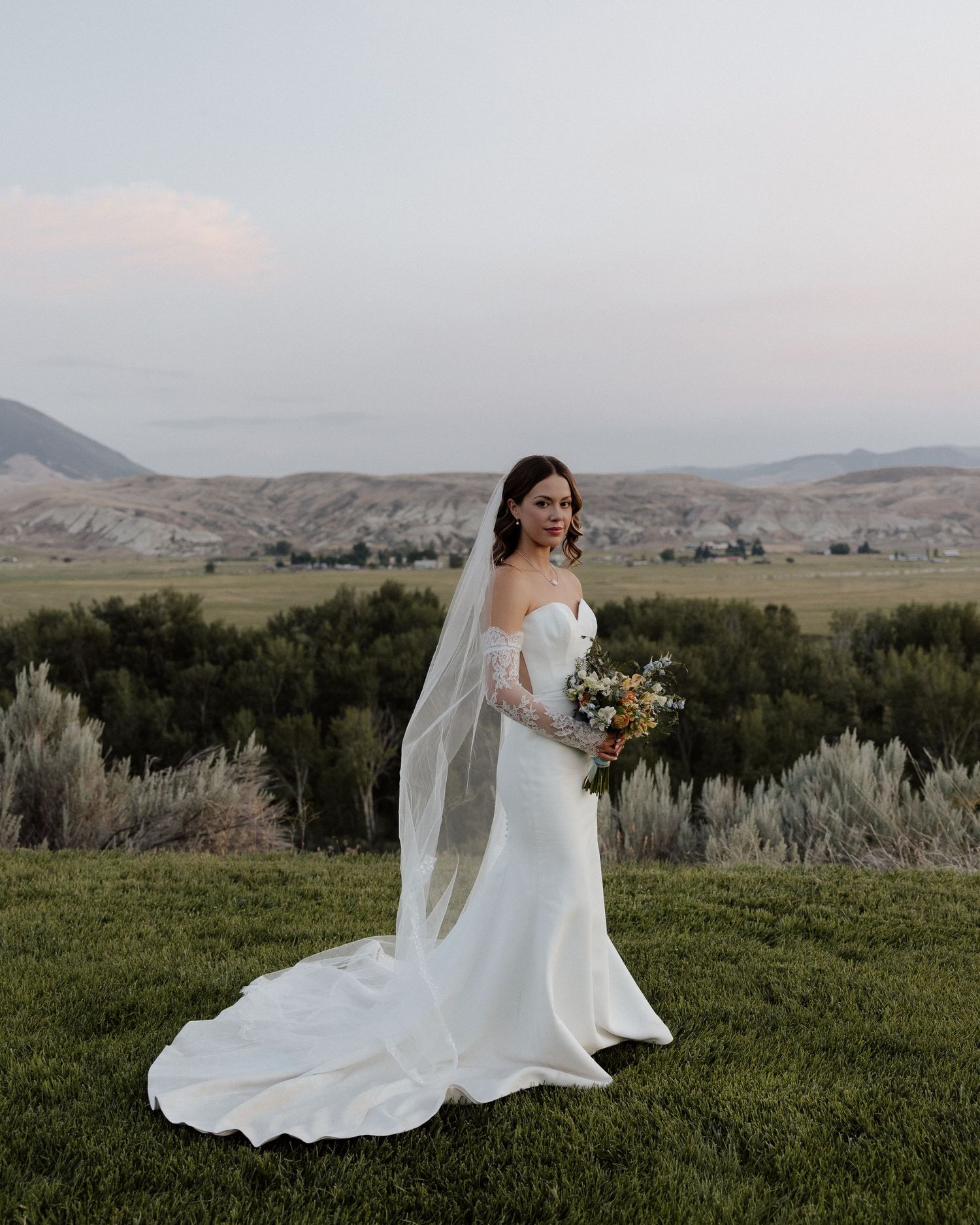 Bride in elegant white dress with train and veil poses in mountain landscape at sunset.