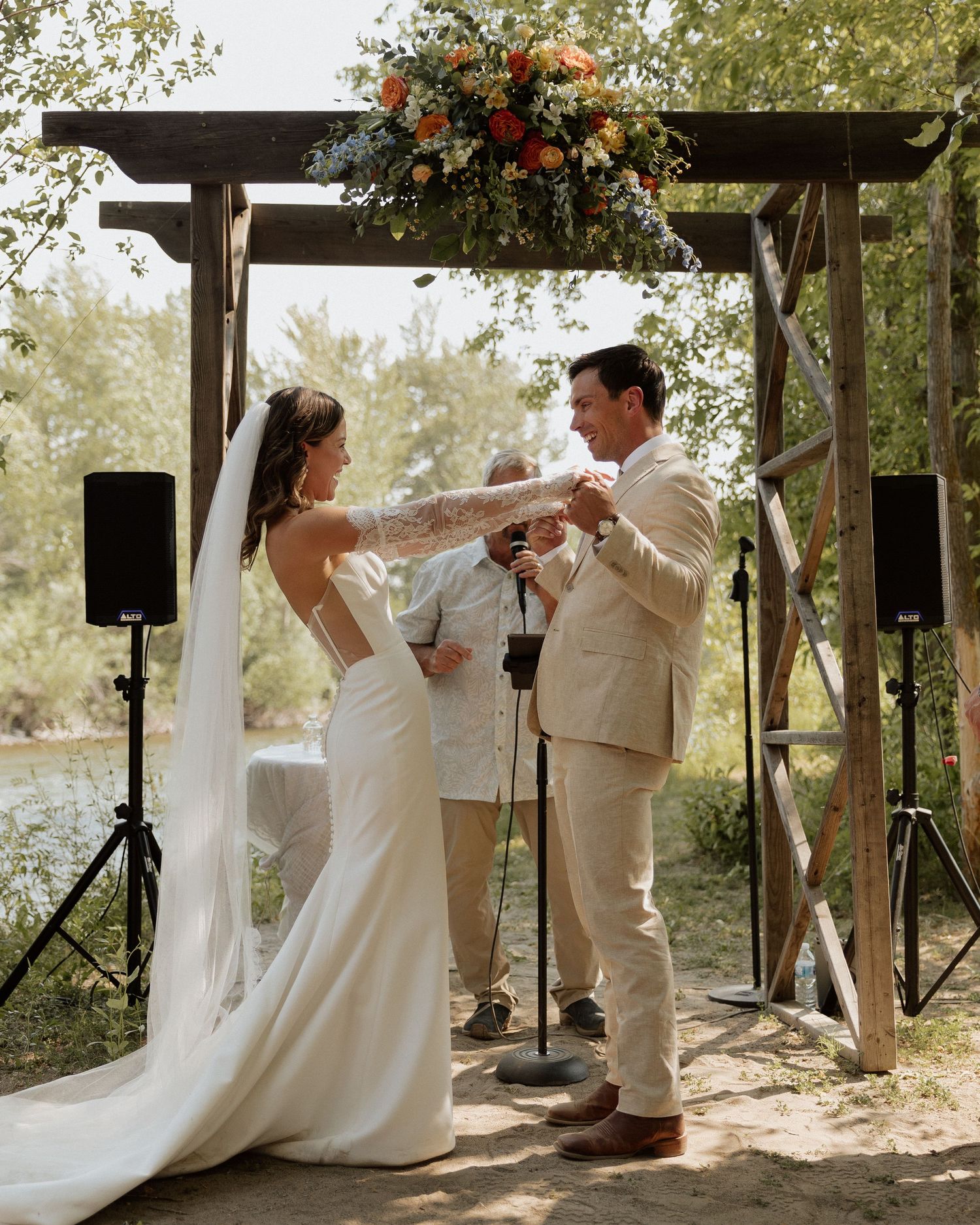 A couple exchanges rings under a wooden arch decorated with flowers during their outdoor wedding ceremony.