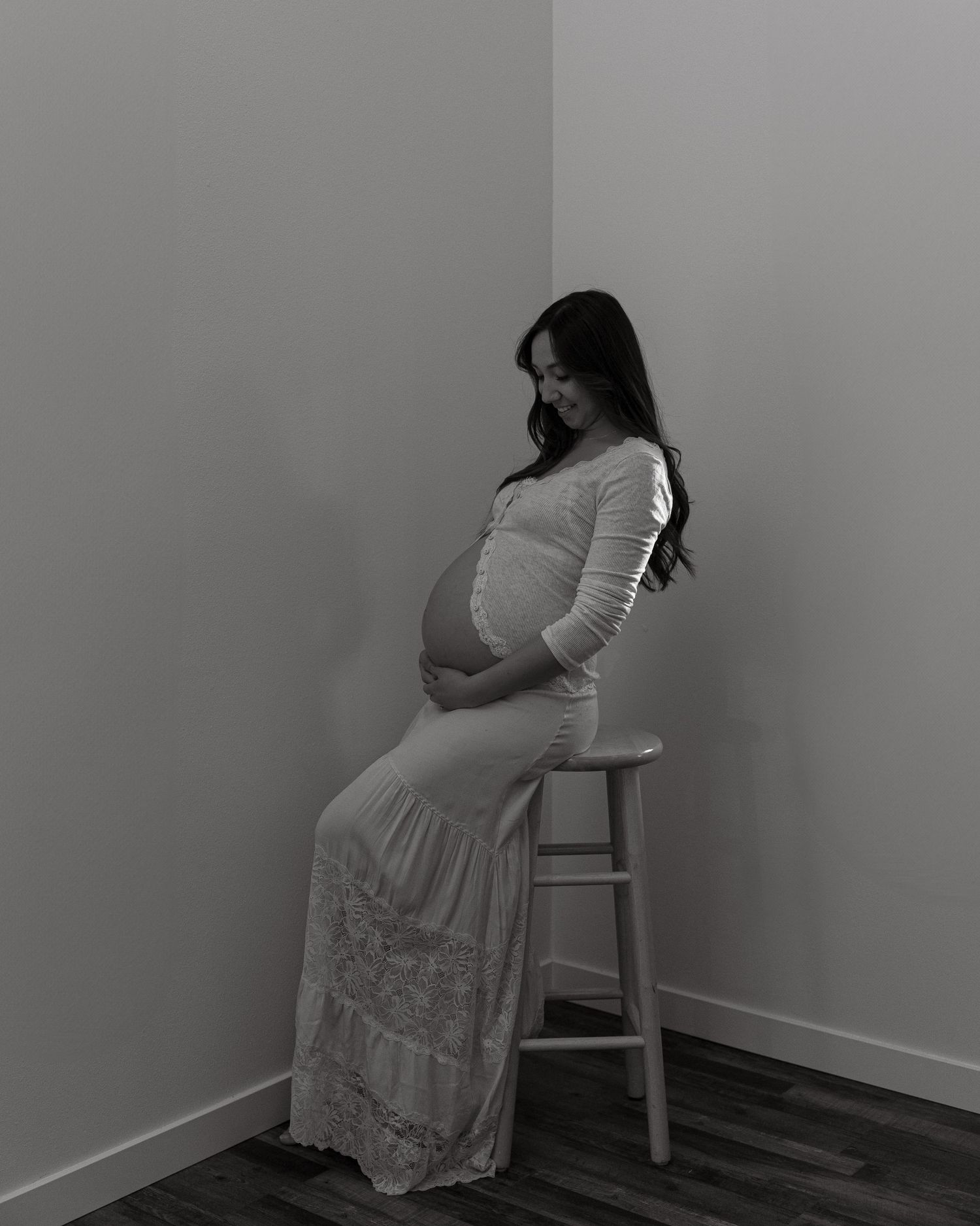 Black and white portrait of an expectant mother in lace dress sitting on wooden stool.