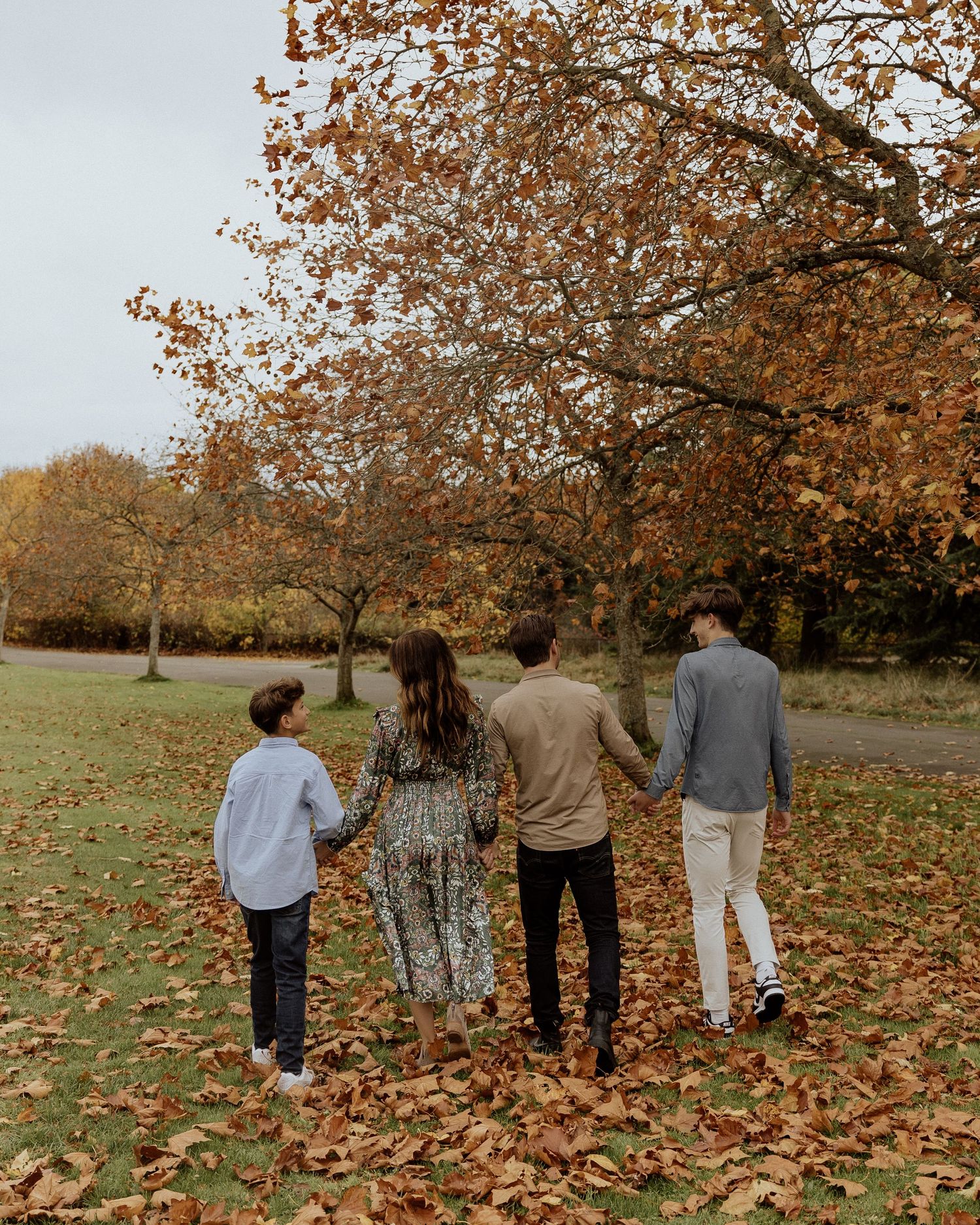 A family walks together through a park filled with autumn trees and fallen leaves.