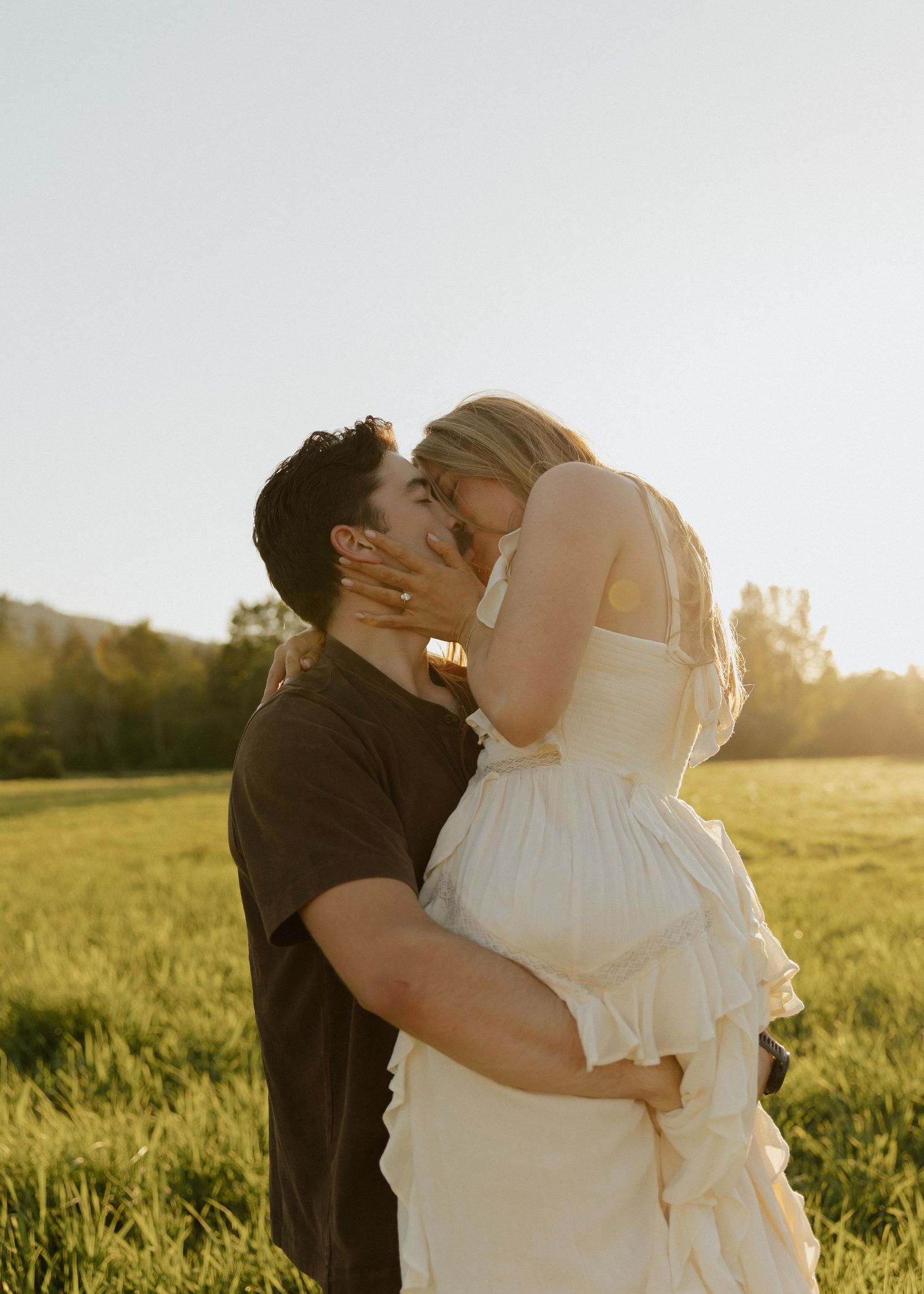 Couple sharing a romantic kiss in a sunlit green field, woman in white ruffled dress, golden hour outdoor engagement photo.