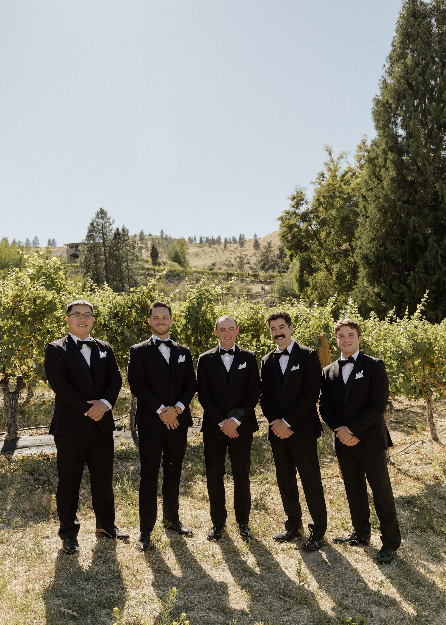 Five groomsmen in black tuxedos pose together at a vineyard wedding venue on a sunny day.
