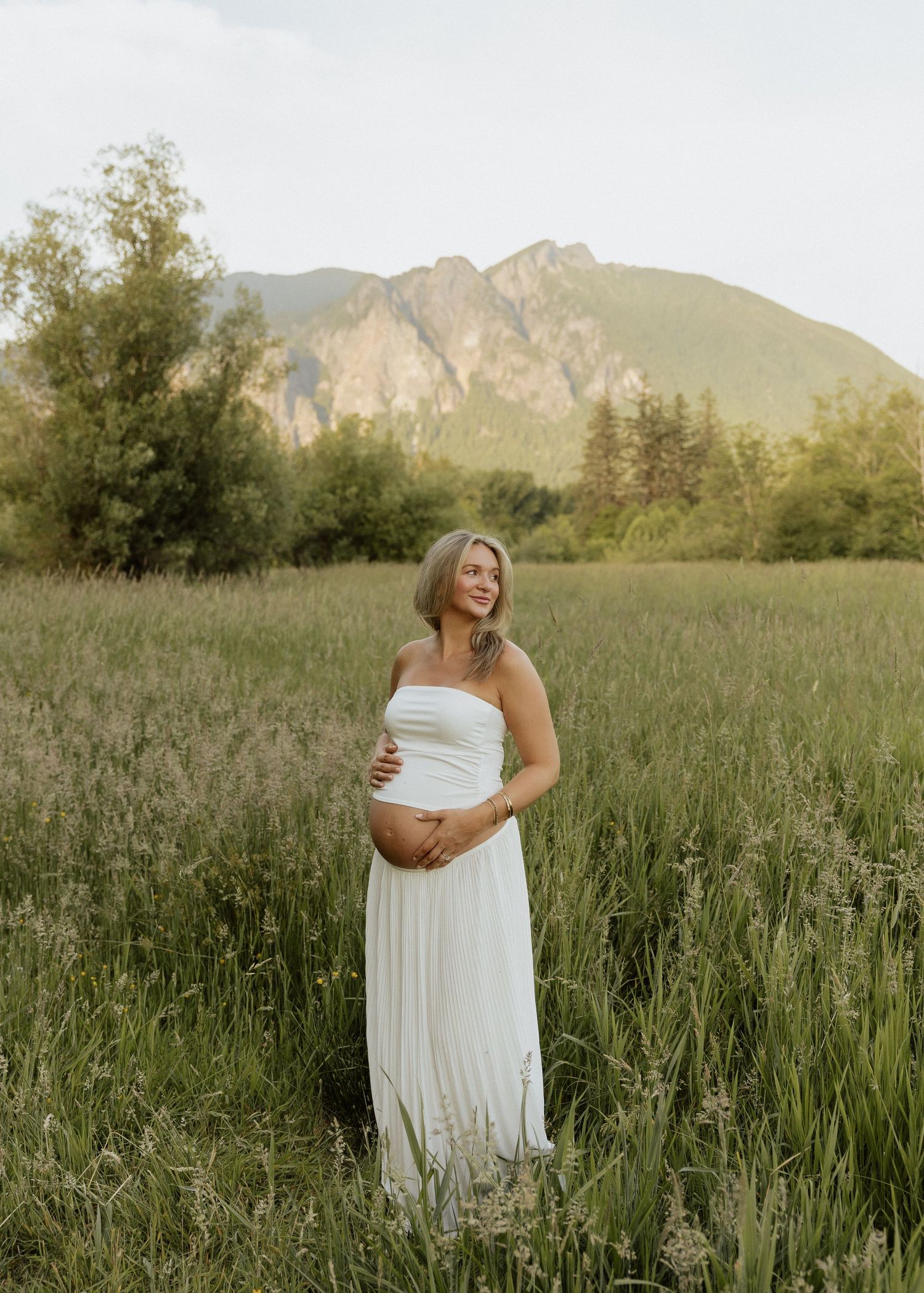 Pregnant woman in white strapless gown standing in lush green meadow with mountain backdrop during golden hour.