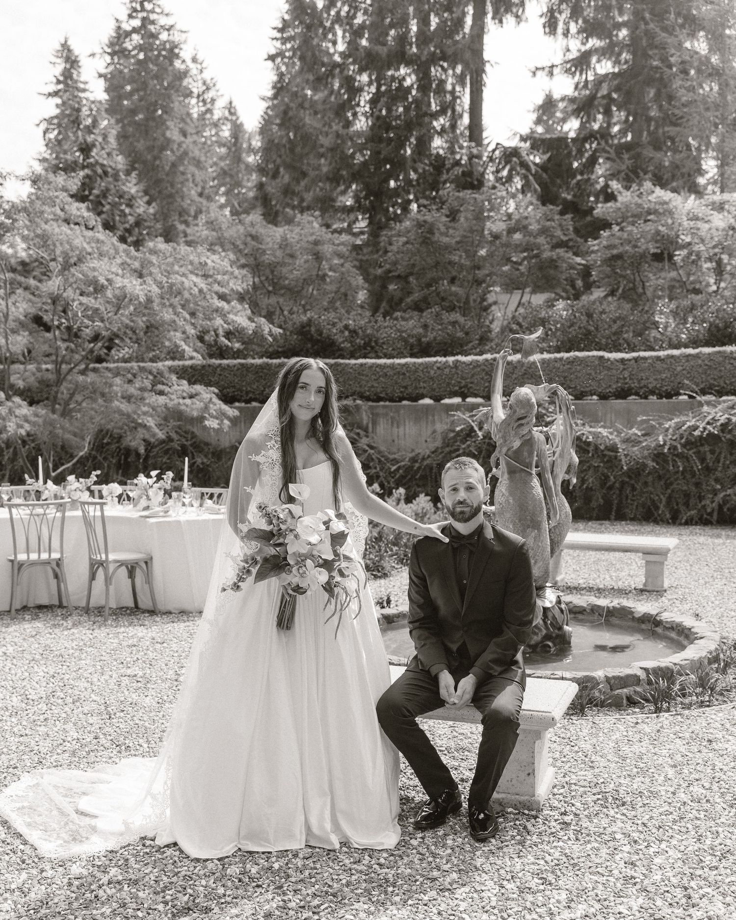Black and white photo of bride and groom posing near fountain with wedding party in lush garden.