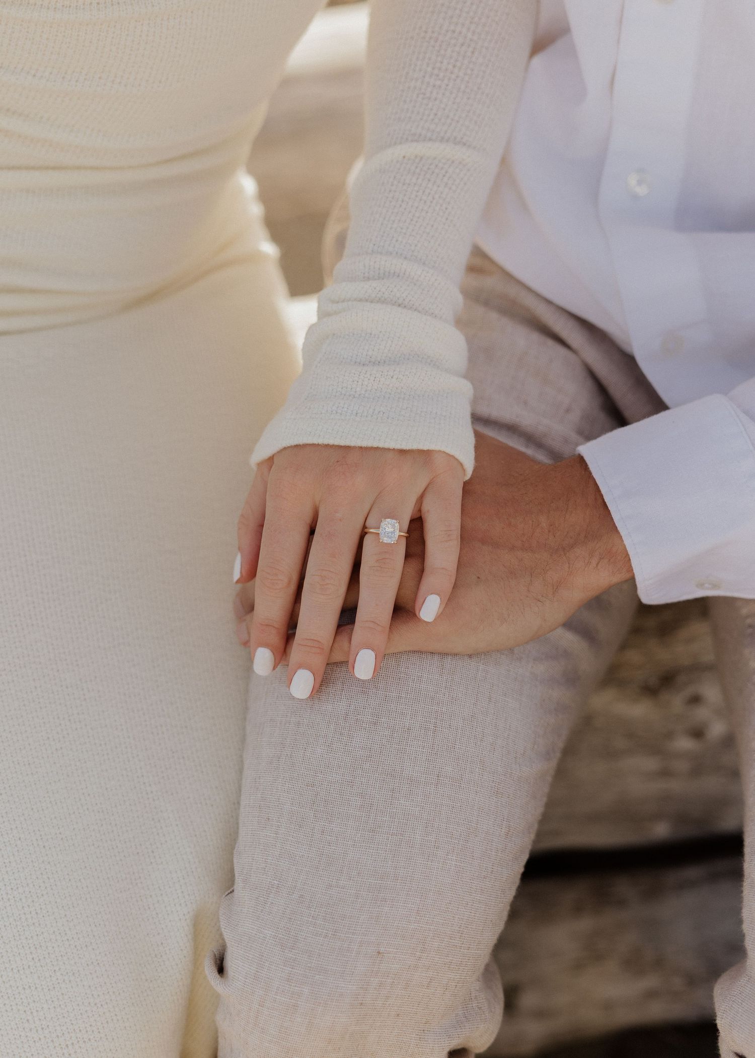 Close-up of a woman's hand wearing a diamond engagement ring resting on a man's knee, both dressed in neutral tones.