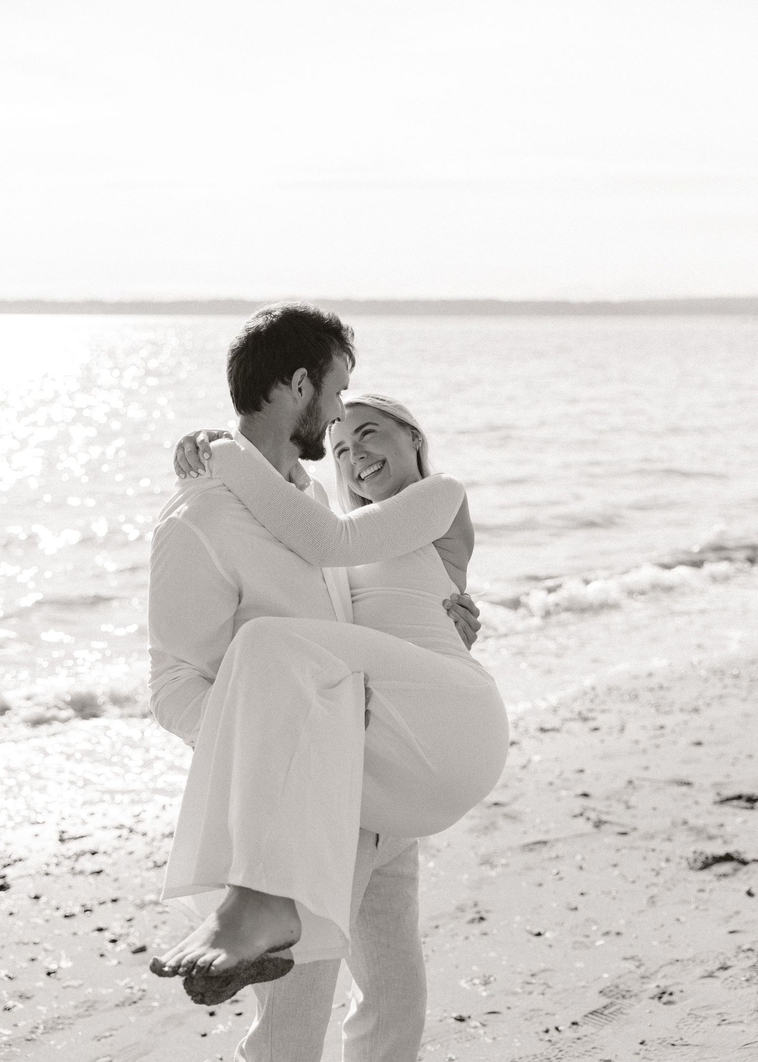 Black and white beach wedding photo of groom carrying bride, both in white, laughing joyfully by the ocean.