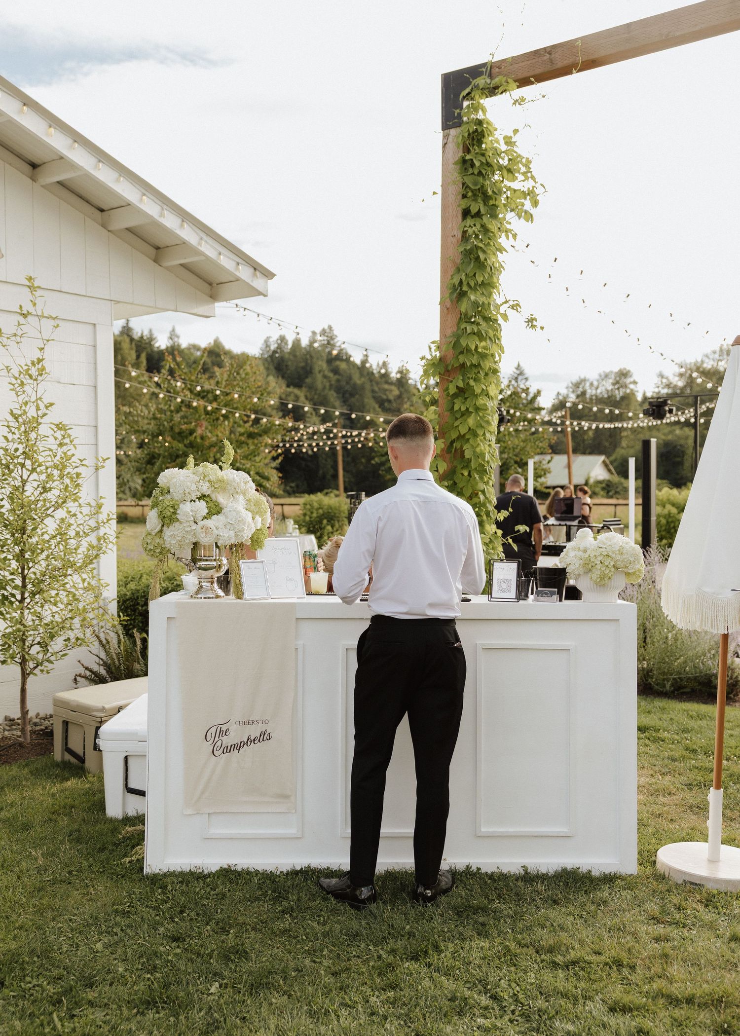 Bartender stands at elegant white outdoor wedding bar with white floral arrangements and string lights at The Campbells reception.