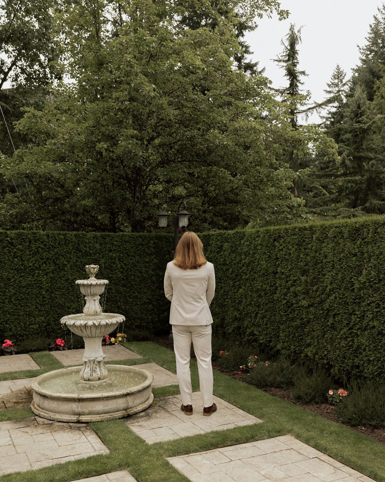 A woman in white outfit stands before a stone fountain in a manicured garden with tall hedges and trees.