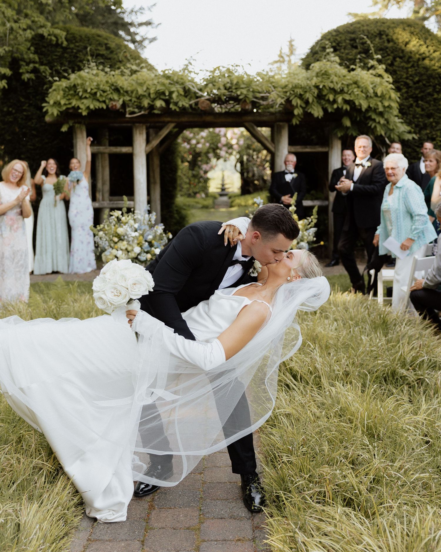Groom dips bride for a kiss after outdoor wedding ceremony, her white veil flowing, guests applauding in background.