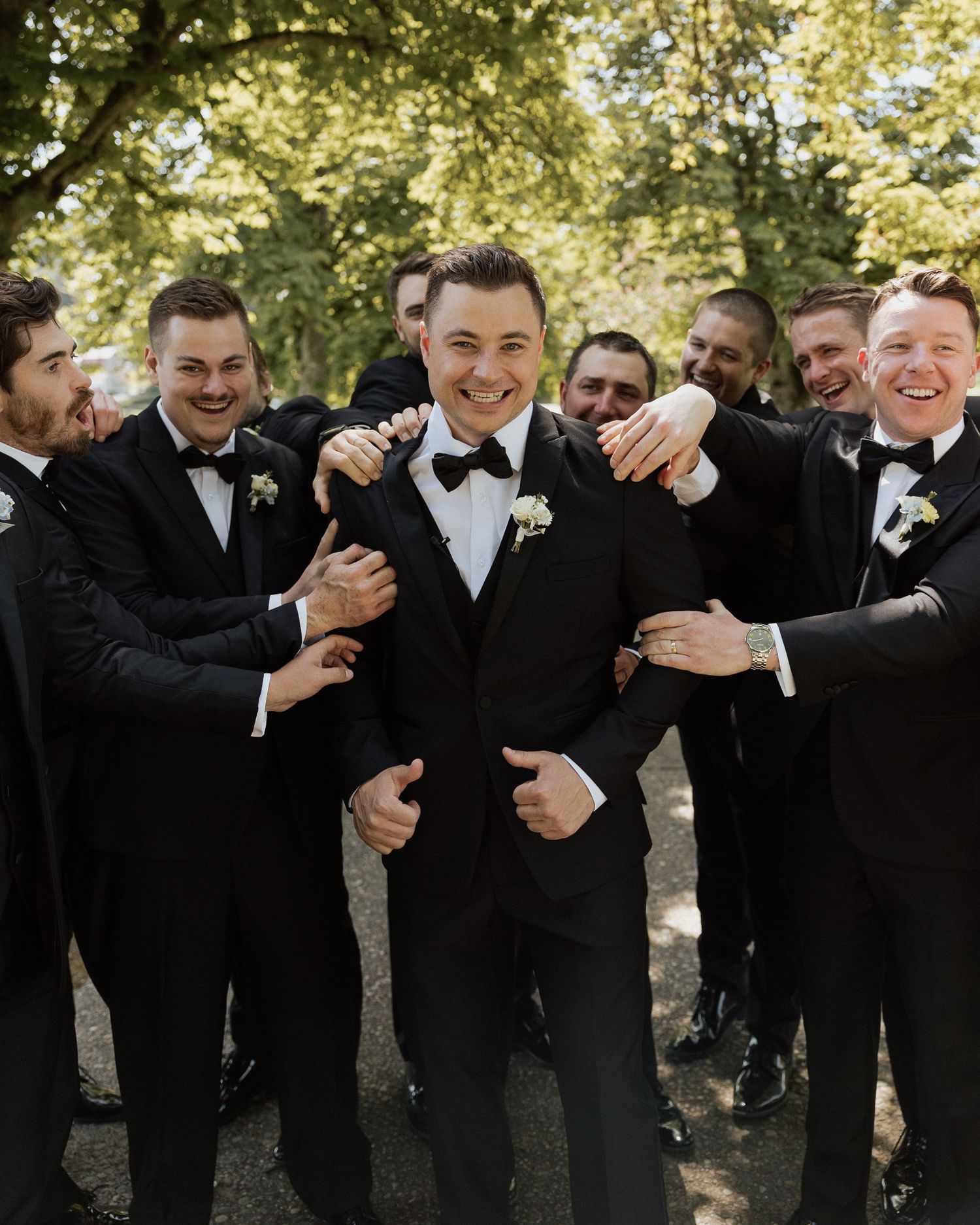 Groom surrounded by smiling groomsmen in black tuxedos with boutonnieres, laughing outdoors at a wedding.