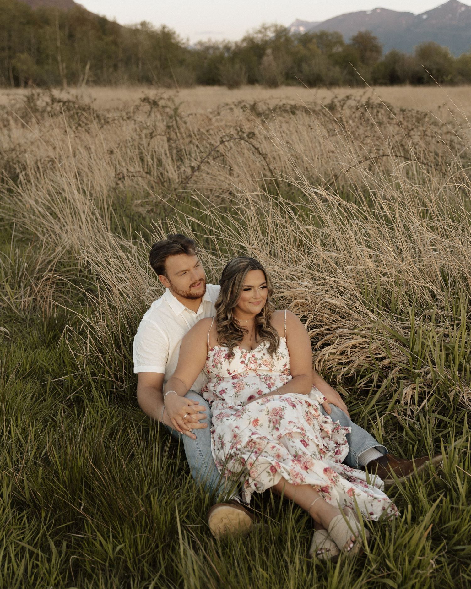 Couple sitting together in tall grass with mountain vista in background.