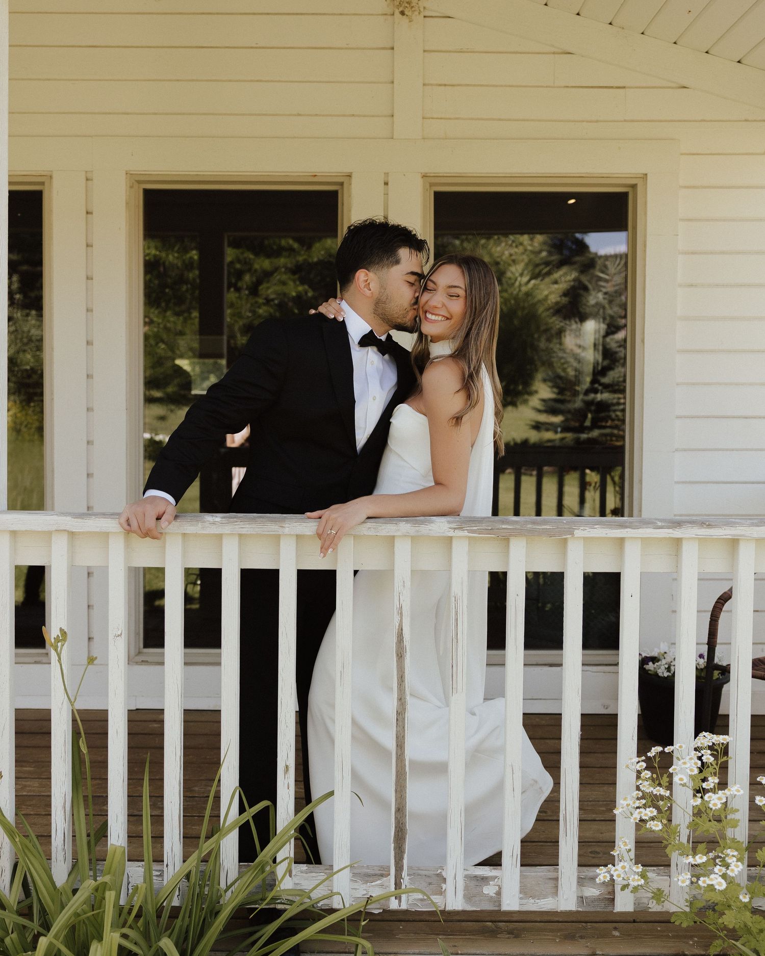A romantic wedding couple embraces on a white porch railing of a house during their special day.