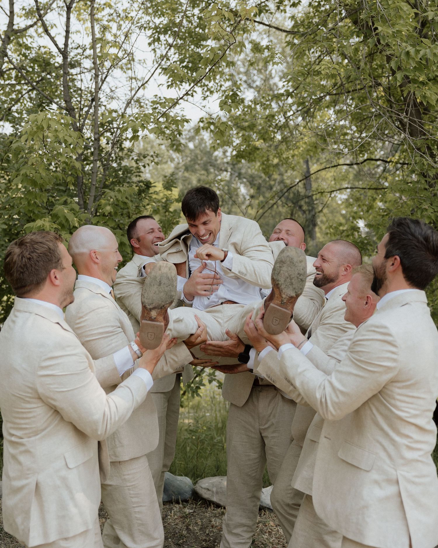 Wedding party lifting and supporting groom in beige suit during outdoor photo session.