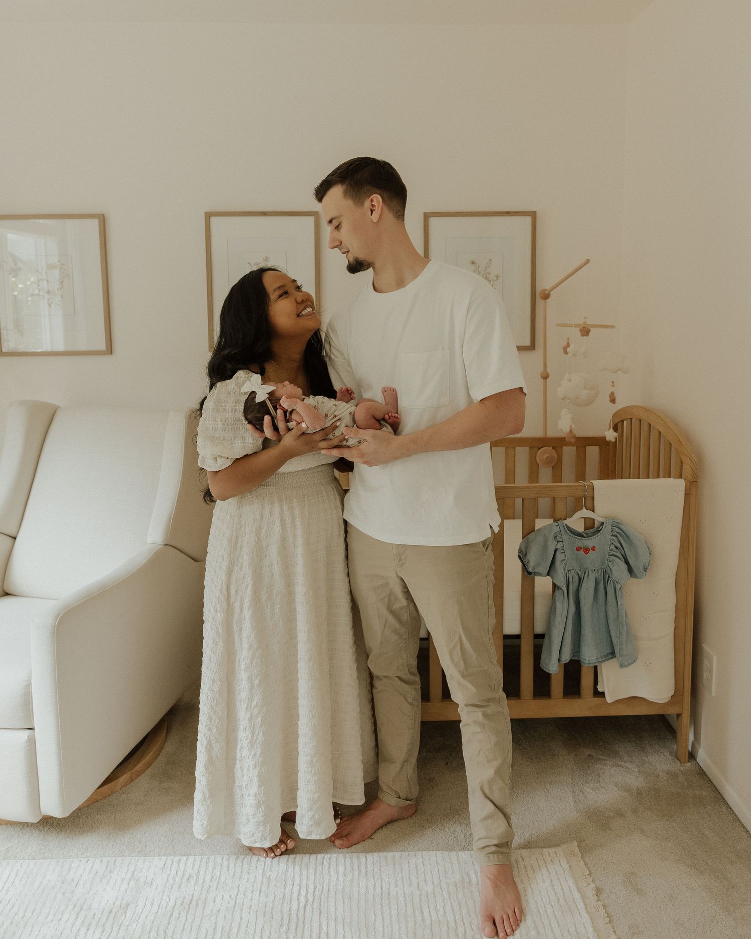 Parents in white and beige outfits tenderly holding their newborn in minimalist nursery setting.