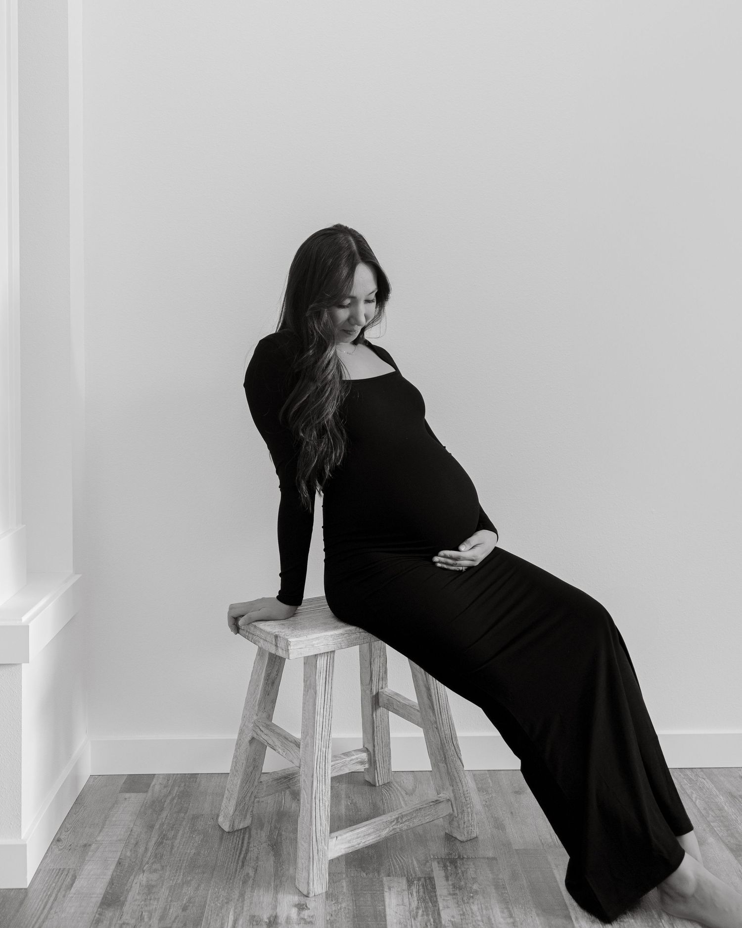 Maternity portrait in black and white showing woman seated on wooden stool in flowing dress.