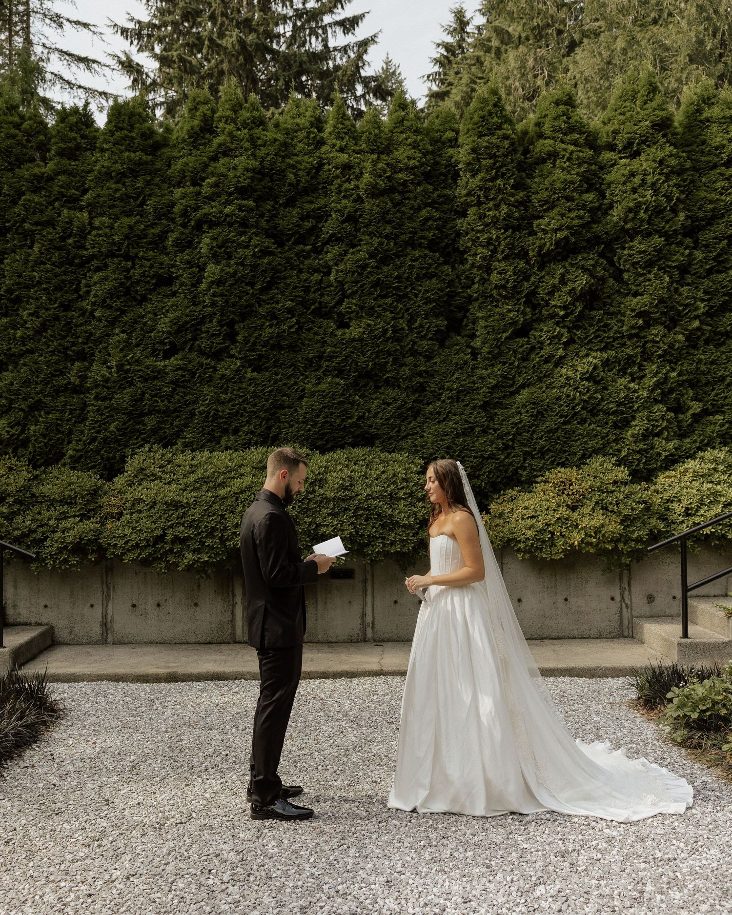 Bride and groom exchange vows outdoors on gravel path with lush green hedge backdrop during intimate wedding ceremony.