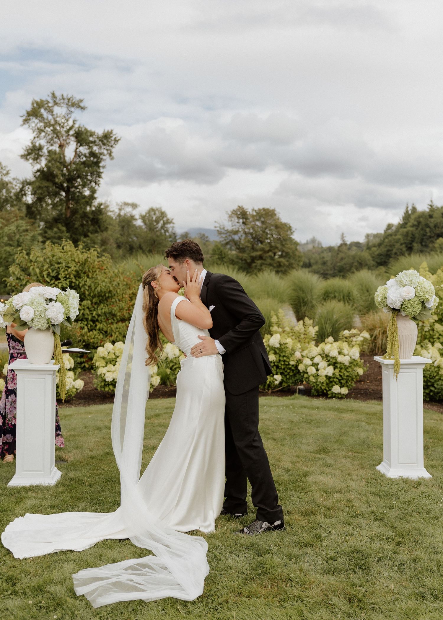 Bride and groom share first kiss at outdoor wedding ceremony with white pillar floral arrangements and lush green garden backdrop.