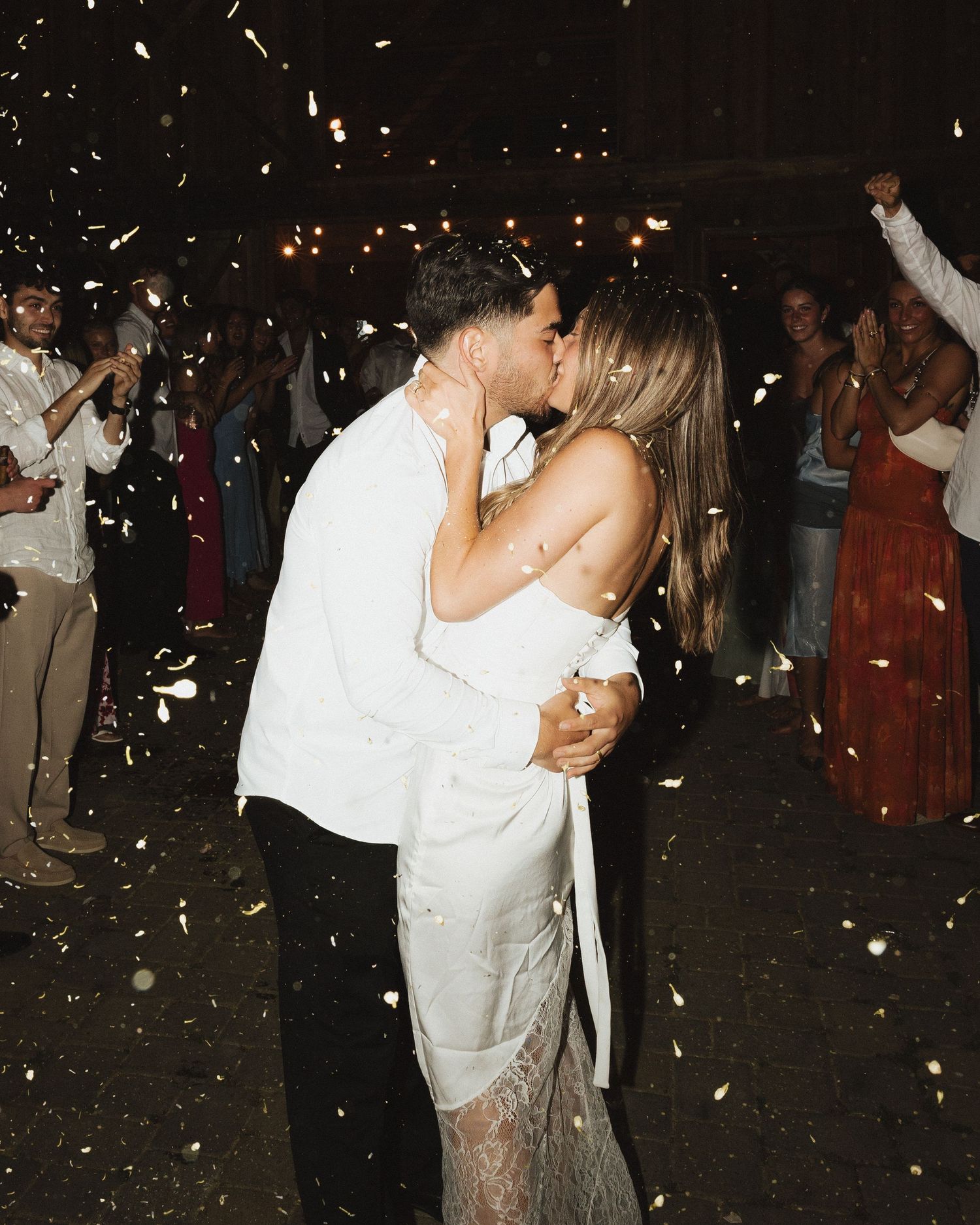 Bride and groom share romantic kiss during grand exit with confetti and sparklers at nighttime wedding reception.