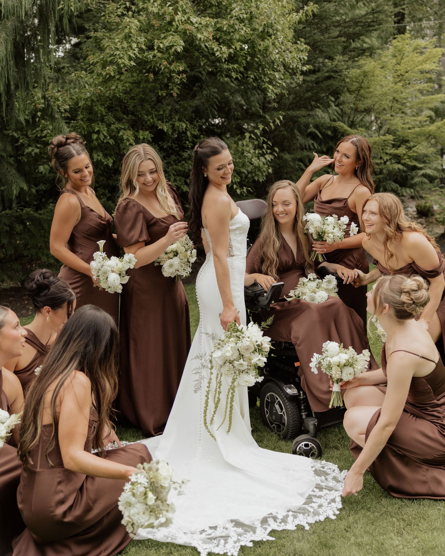 Bride in white gown poses with bridesmaids in brown satin dresses holding white bouquets outdoors.