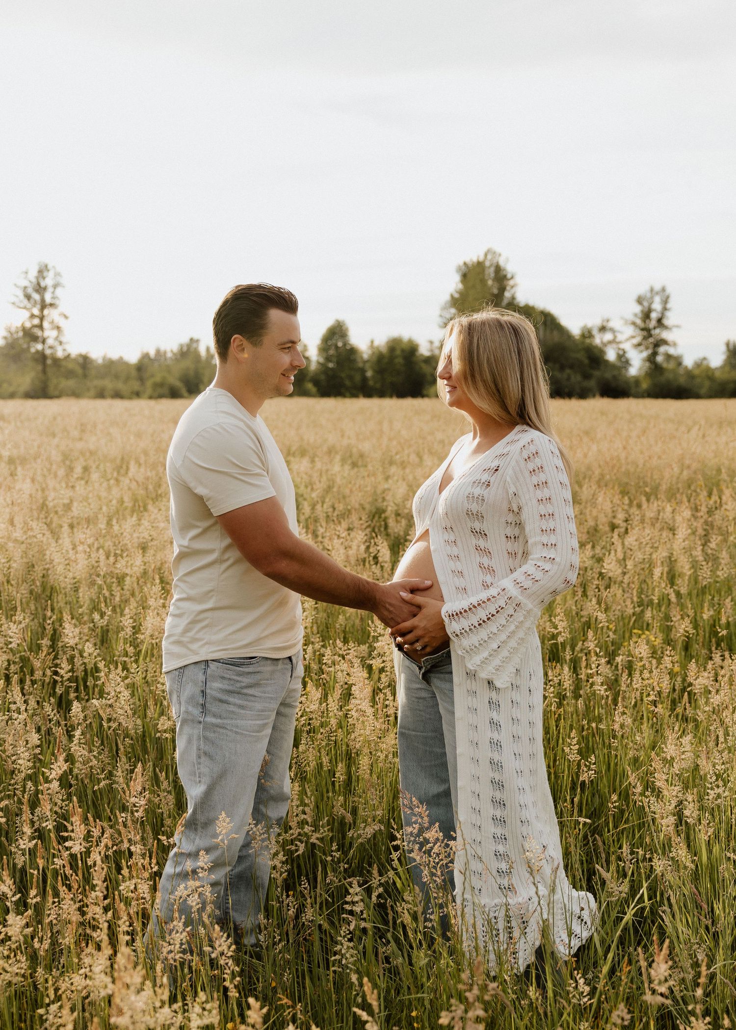 Couple holding hands in a golden field, woman in white lace maternity dress, man in white tee and jeans, trees in background.