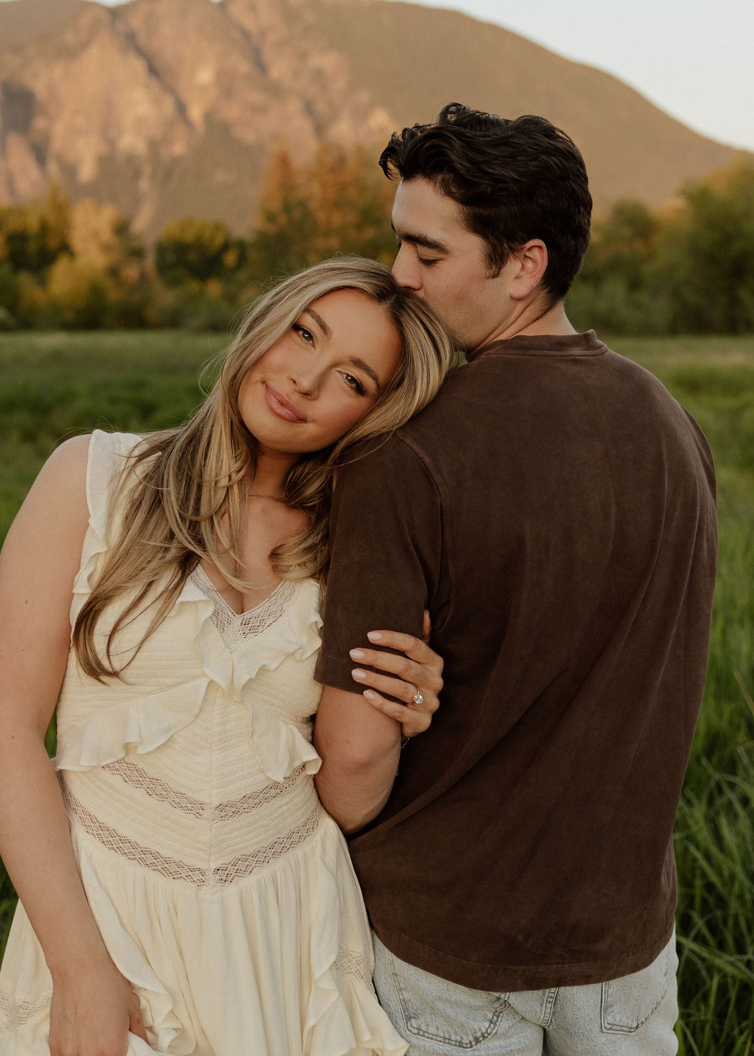 A woman leans her head on her partners shoulder in a field with a mountain backdrop at dusk.