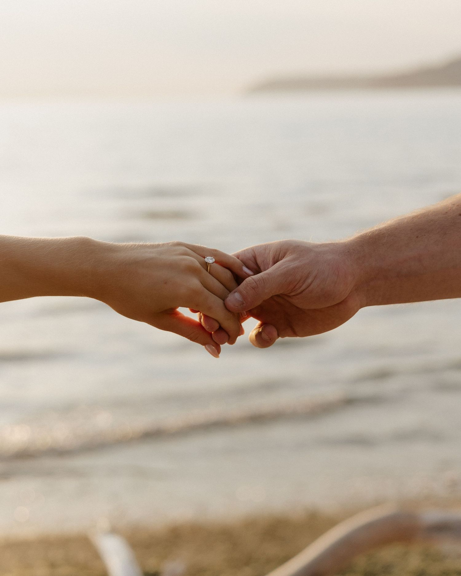 Two hands tenderly clasped together against a sunset beach backdrop.