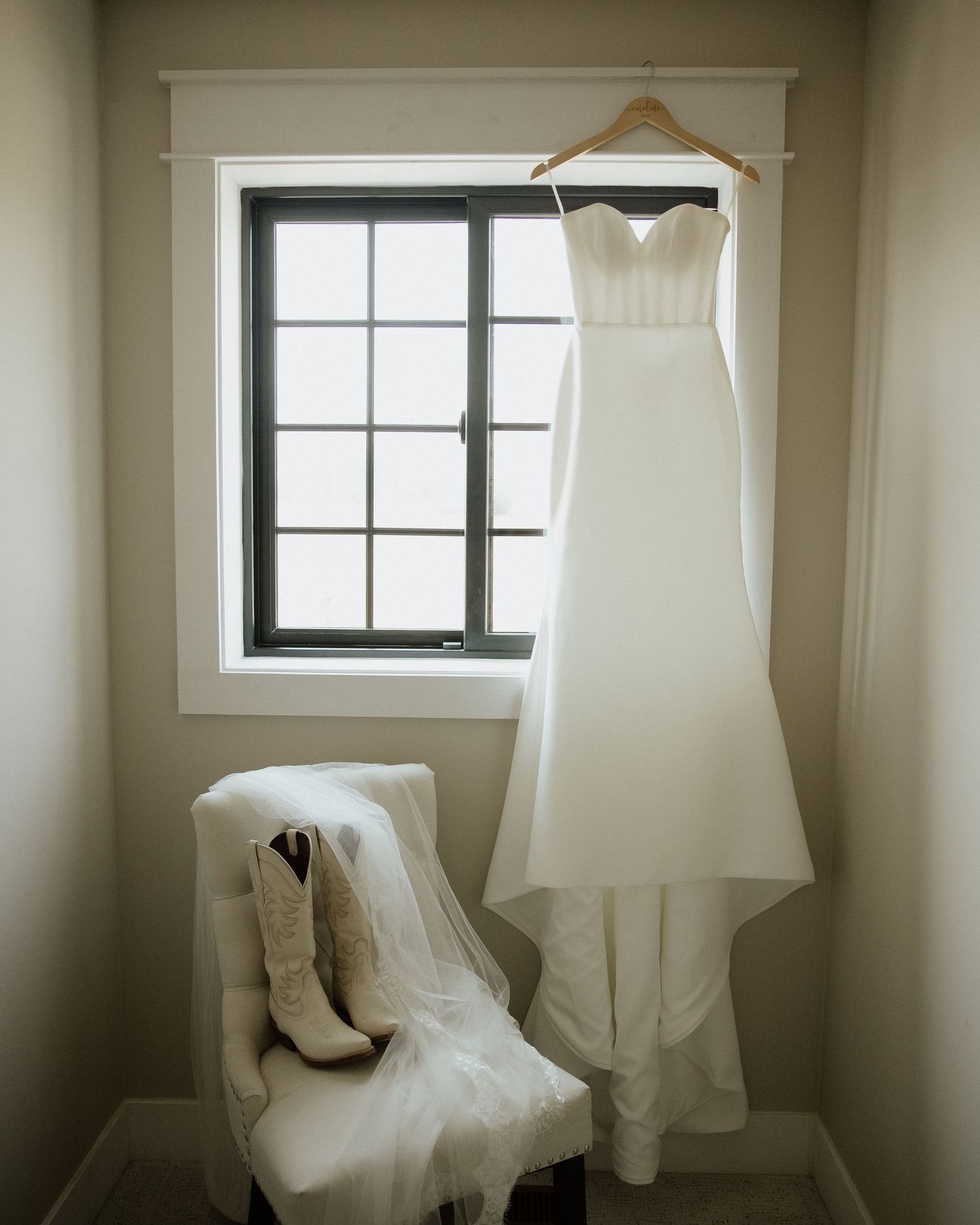 An elegant white wedding dress hangs by a window with cowboy boots placed on a chair below.