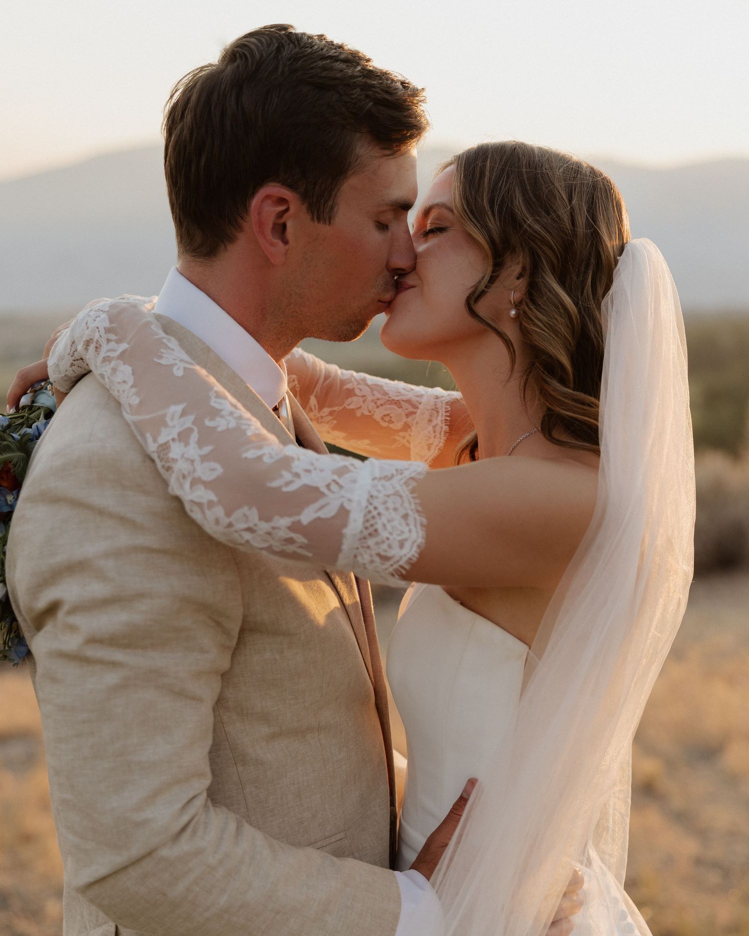 Newlyweds share an intimate kiss during sunset with mountains in the background.