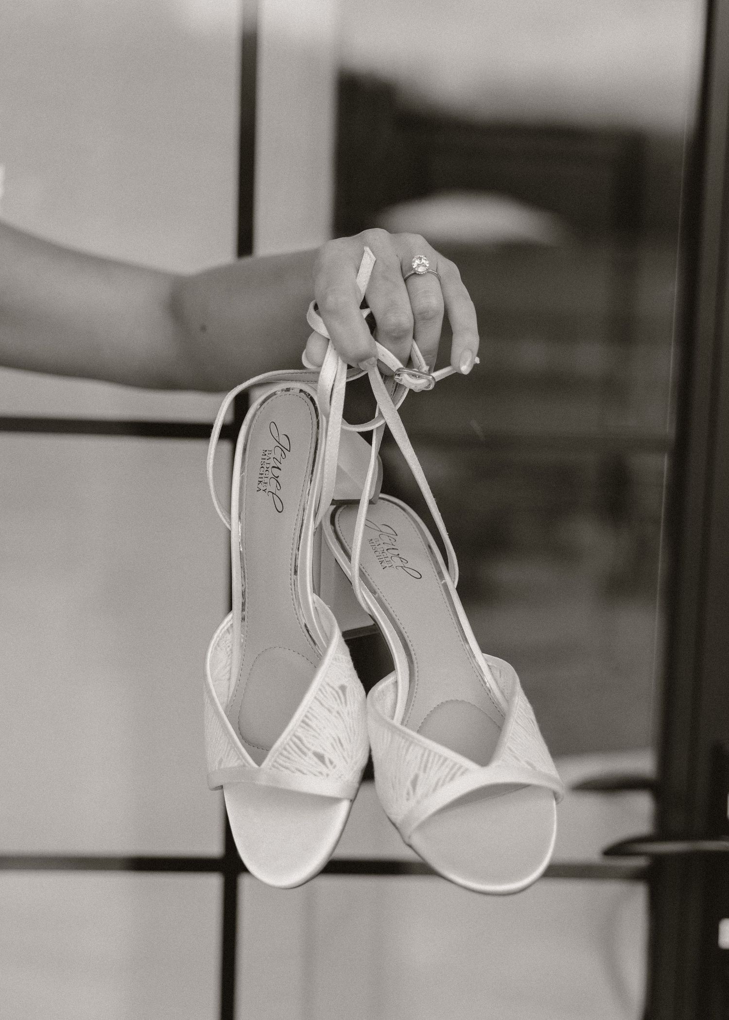 A bride holds elegant white strappy heeled sandals by their ribbons in a black and white wedding photo.