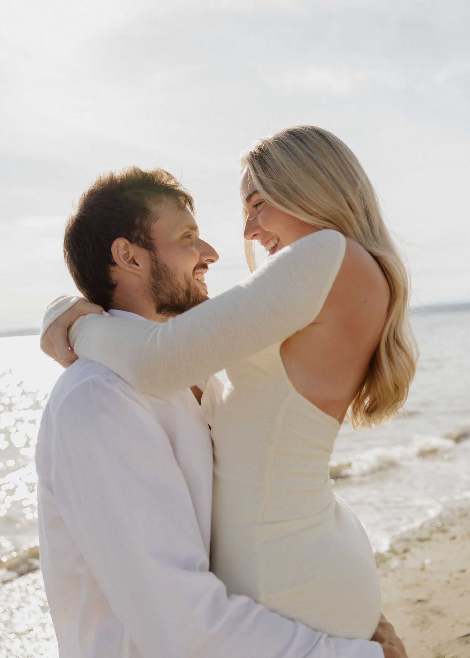 Happy couple embracing and laughing together on a bright sunny beach, woman in backless white dress.