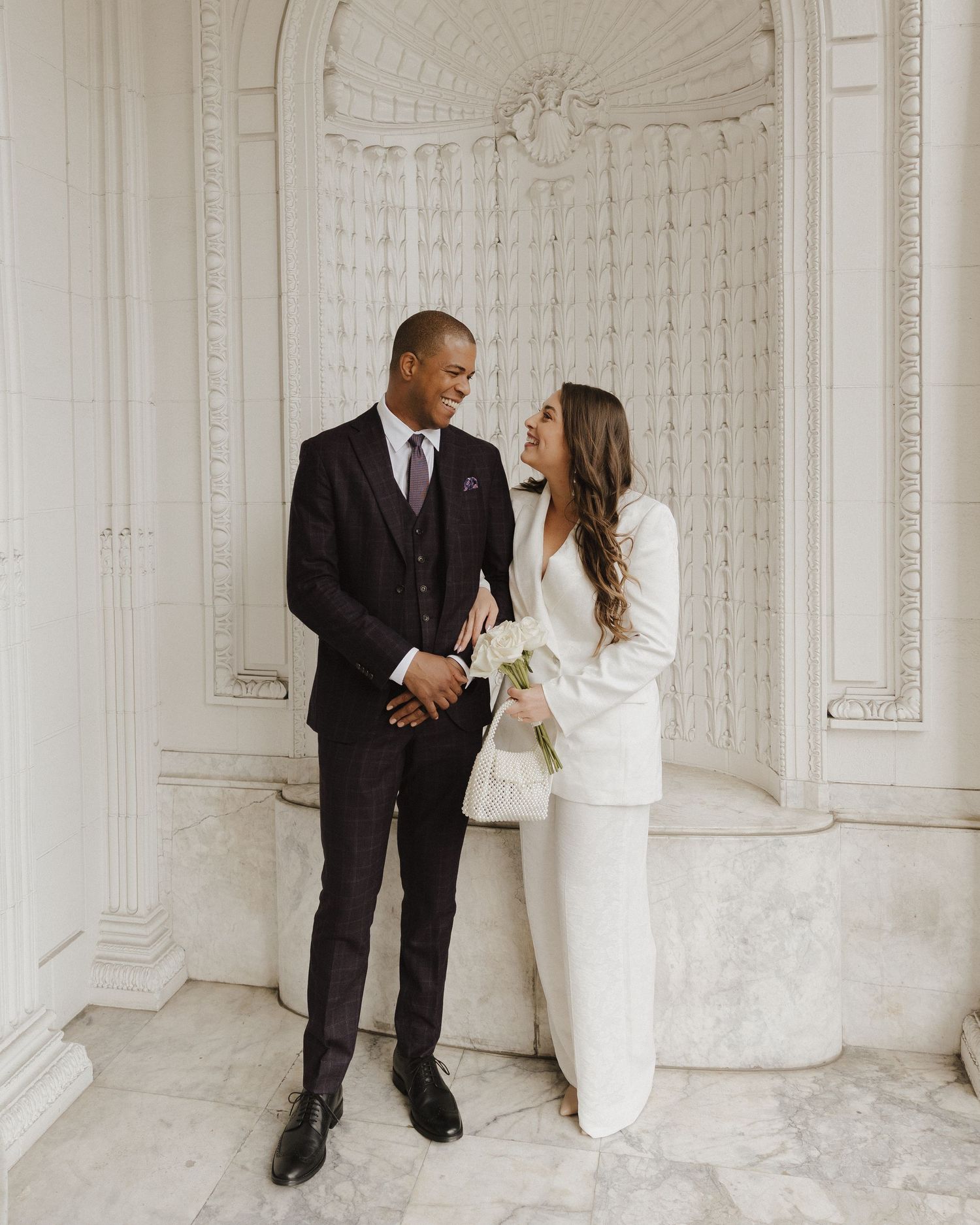 A couple in wedding attire smiles at each other in an elegant marble hall, groom in dark suit, bride in white pantsuit holding flowers.