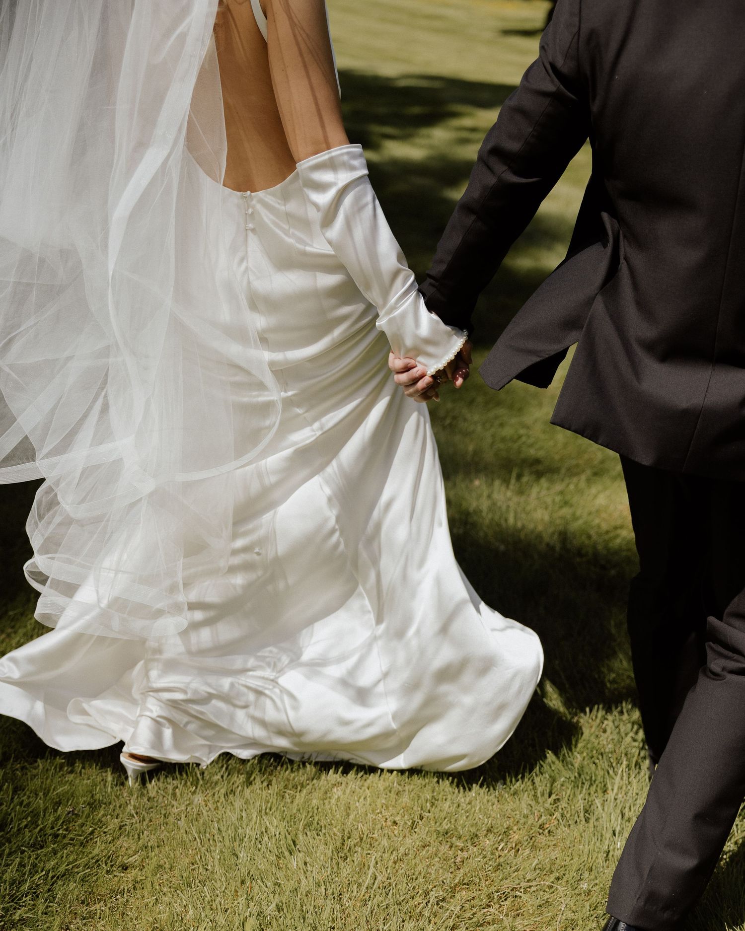 Bride in flowing white satin gown holds hands with groom in dark suit walking on grass.