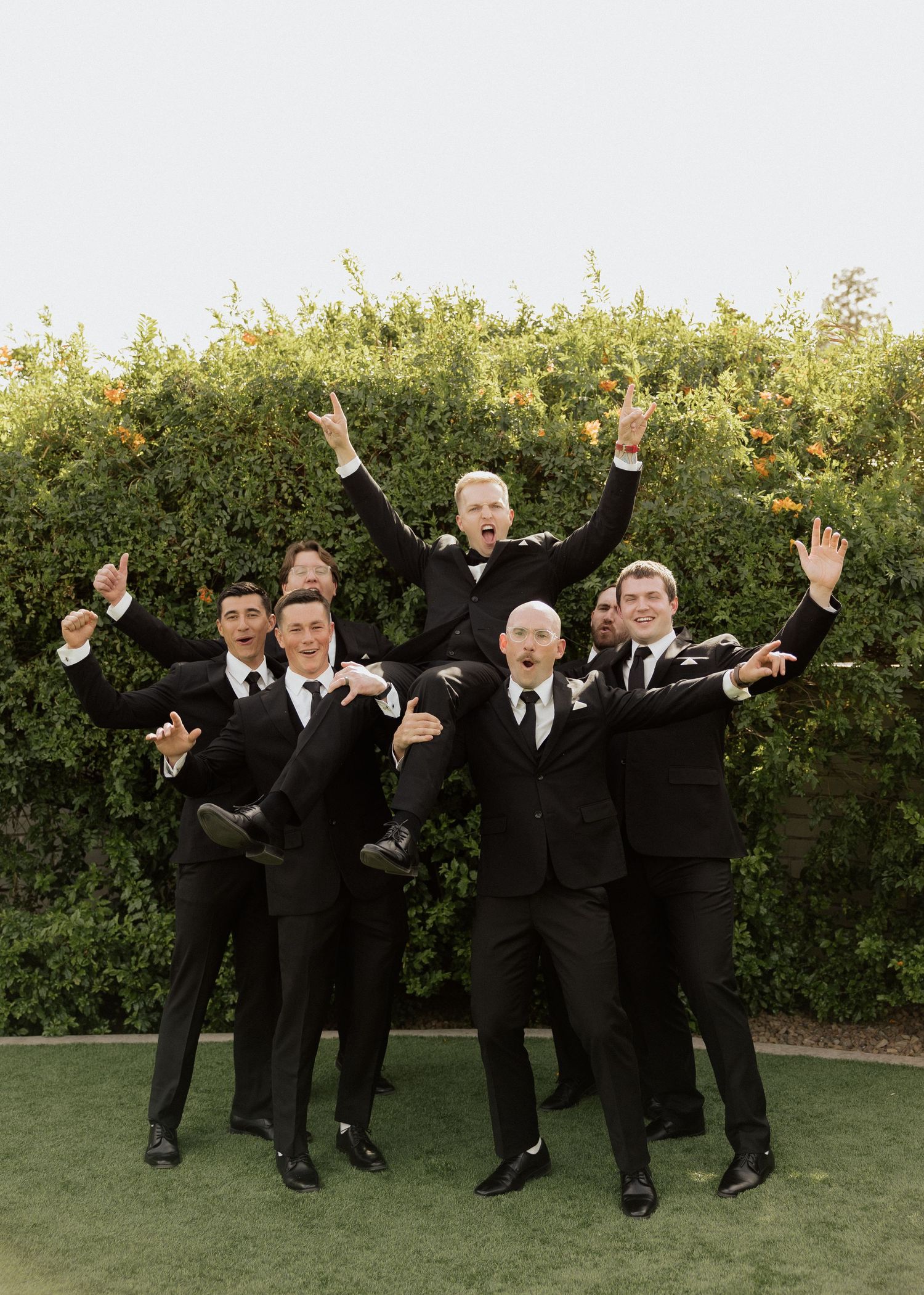 Groomsmen in black tuxedos celebrate outdoors, lifting groom with joyful poses in front of lush green hedges.
