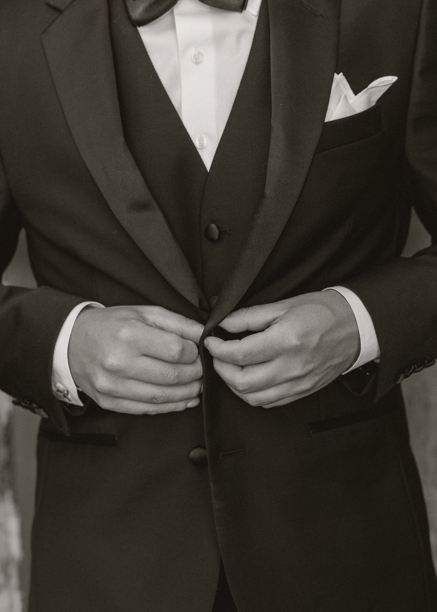 Groom in black tuxedo with bow tie and pocket square buttoning his jacket before wedding ceremony.