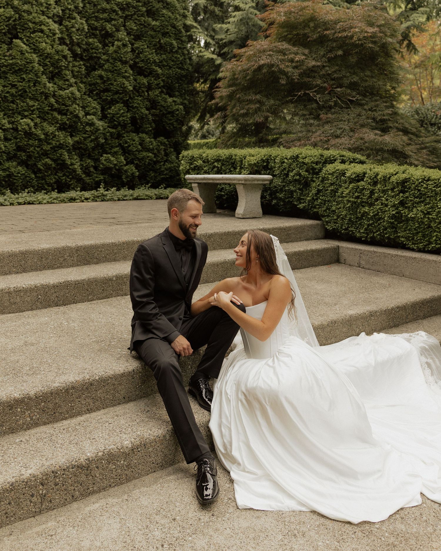 A bride and groom sit together on stone steps in a lush garden, sharing a tender moment on their wedding day.