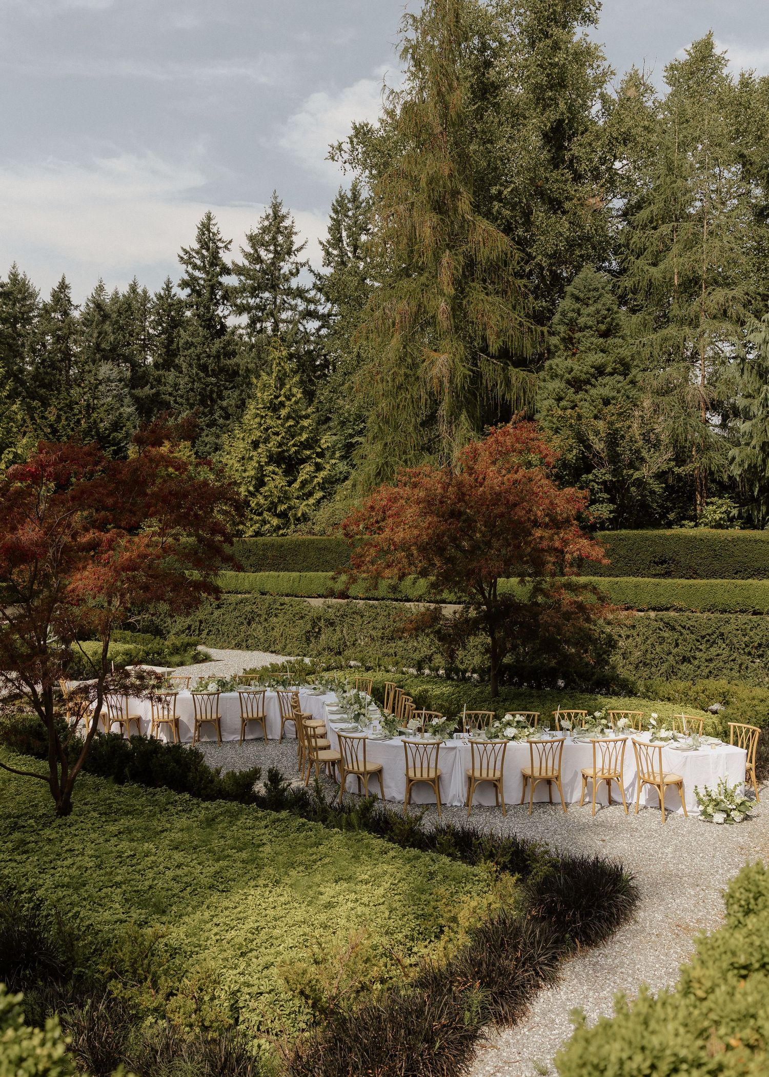 Elegant outdoor wedding reception with long white tables, gold chairs, surrounded by lush greenery and colorful autumn trees.