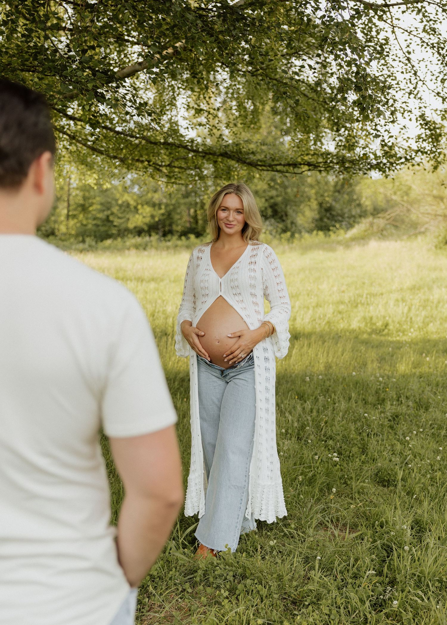 Pregnant woman in white lace cardigan and jeans cradling baby bump in sunny green field while partner looks on.