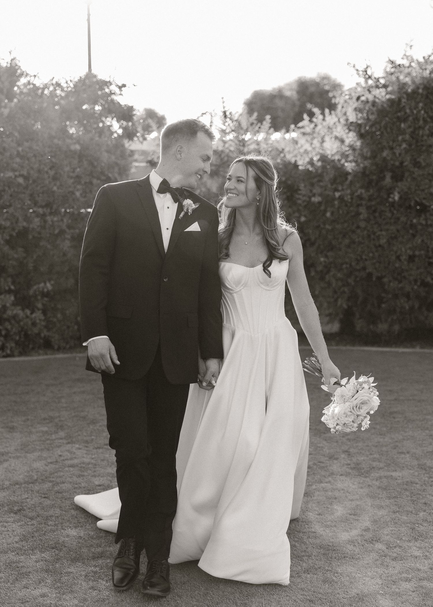 Black and white wedding photo of a smiling bride in strapless gown and groom in tuxedo walking hand in hand outdoors.