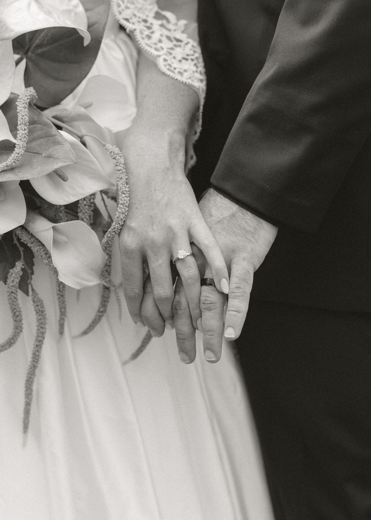 Black and white close-up of newlyweds holding hands showing wedding rings with bridal bouquet of calla lilies.