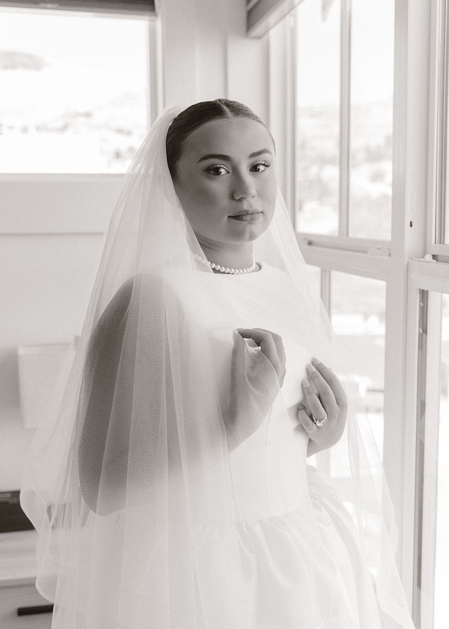 Black and white portrait of a bride in a white gown, veil, and pearl necklace standing by a window.