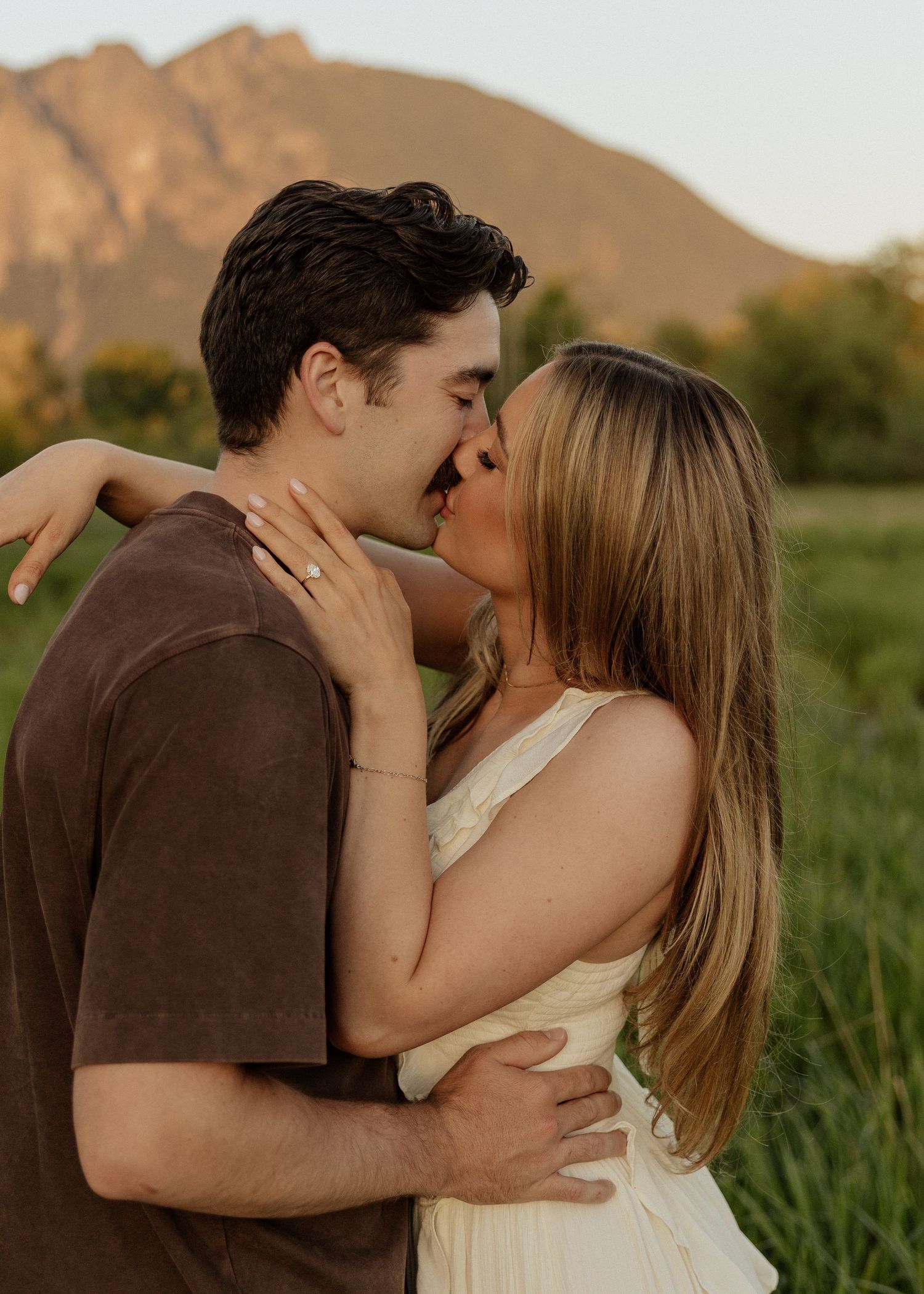 An engaged couple shares a romantic kiss in a green field with a mountain glowing behind them.
