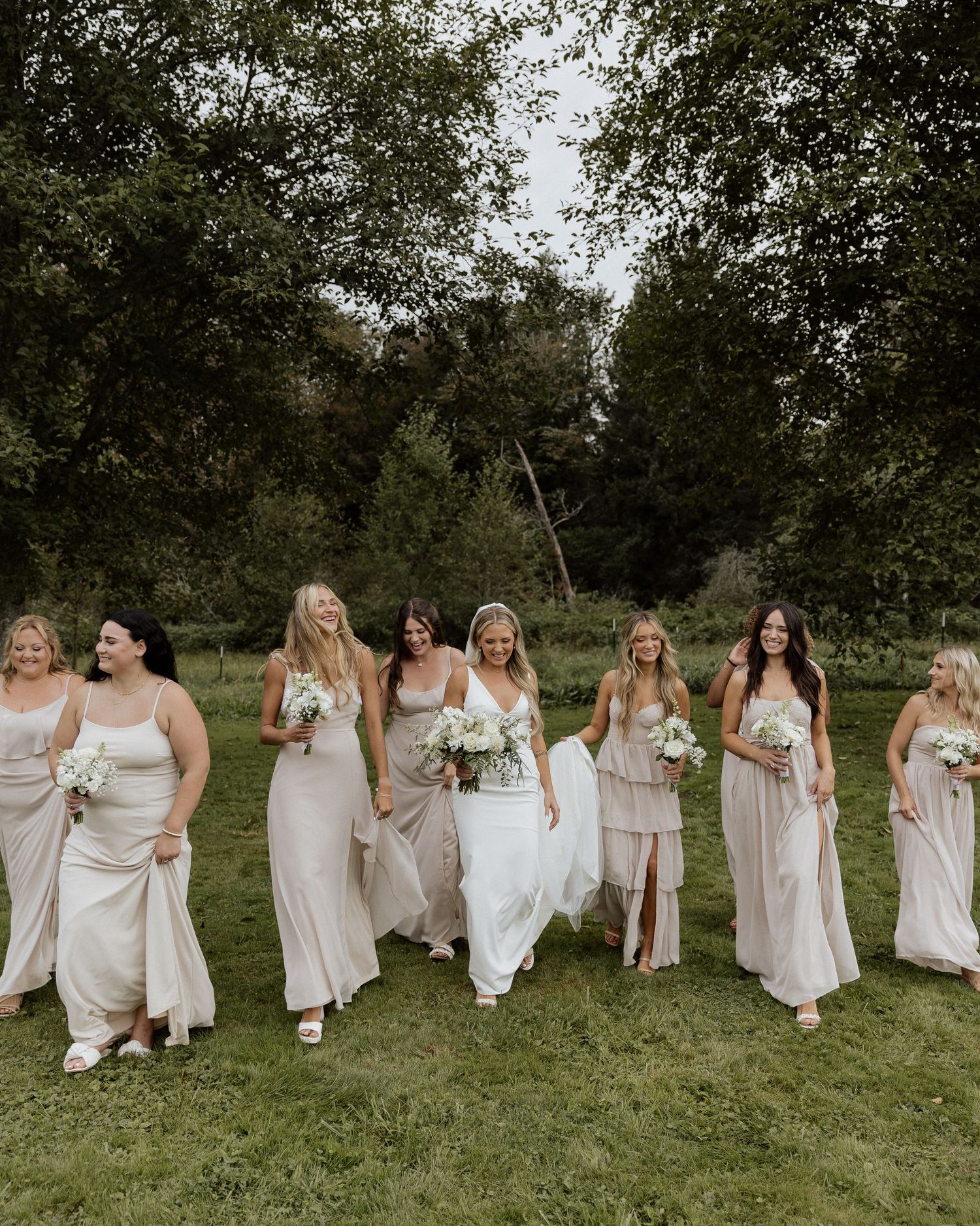 Bridal party wearing neutral dresses walks together on grass laughing and smiling.