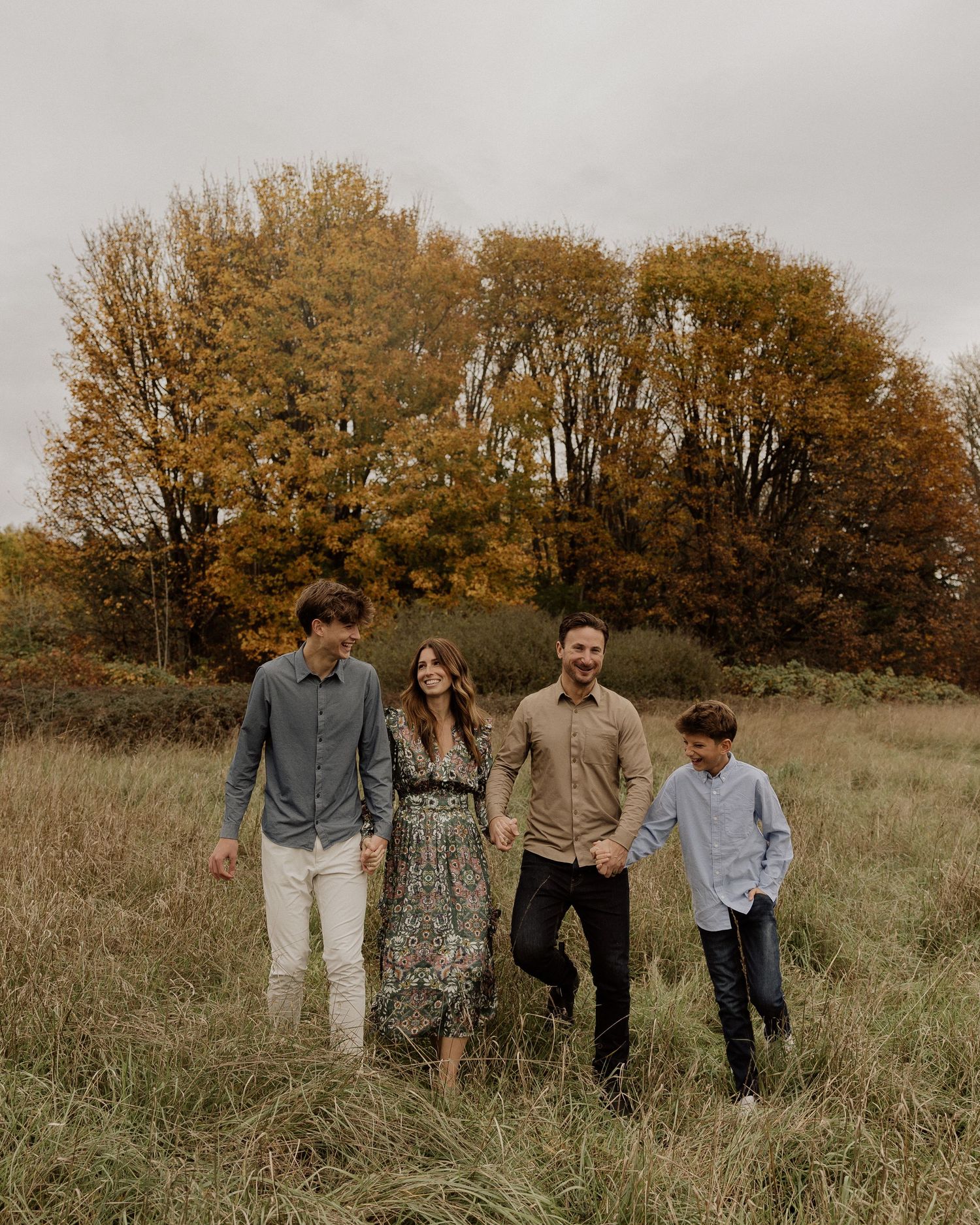Four people walking through tall grass with autumn foliage in background.