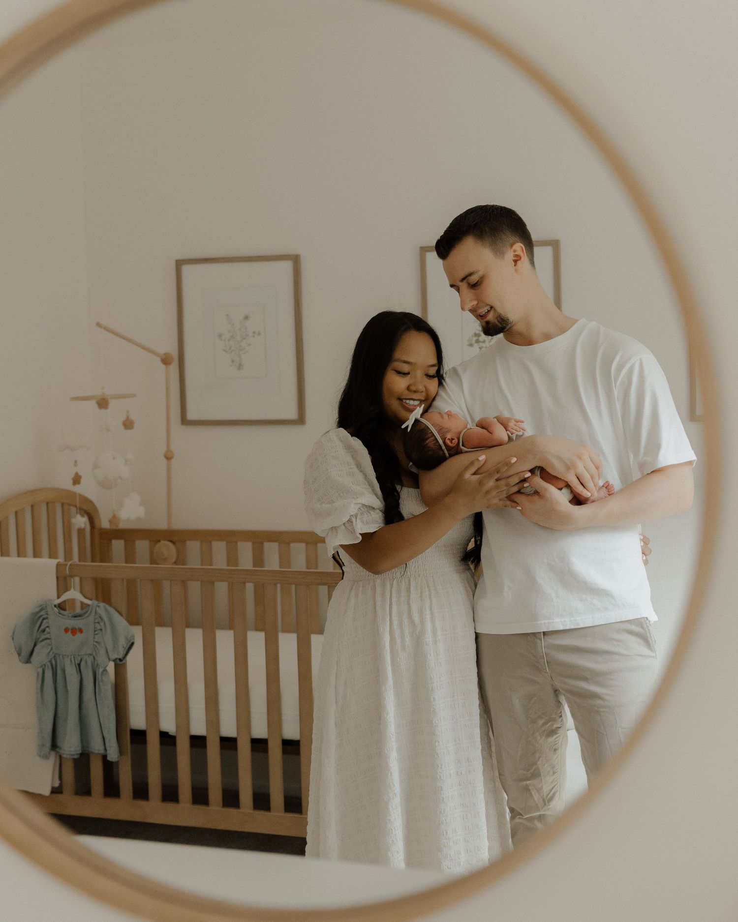 Mirror reflection shows parents admiring their newborn in a cozy nursery with a wooden crib.