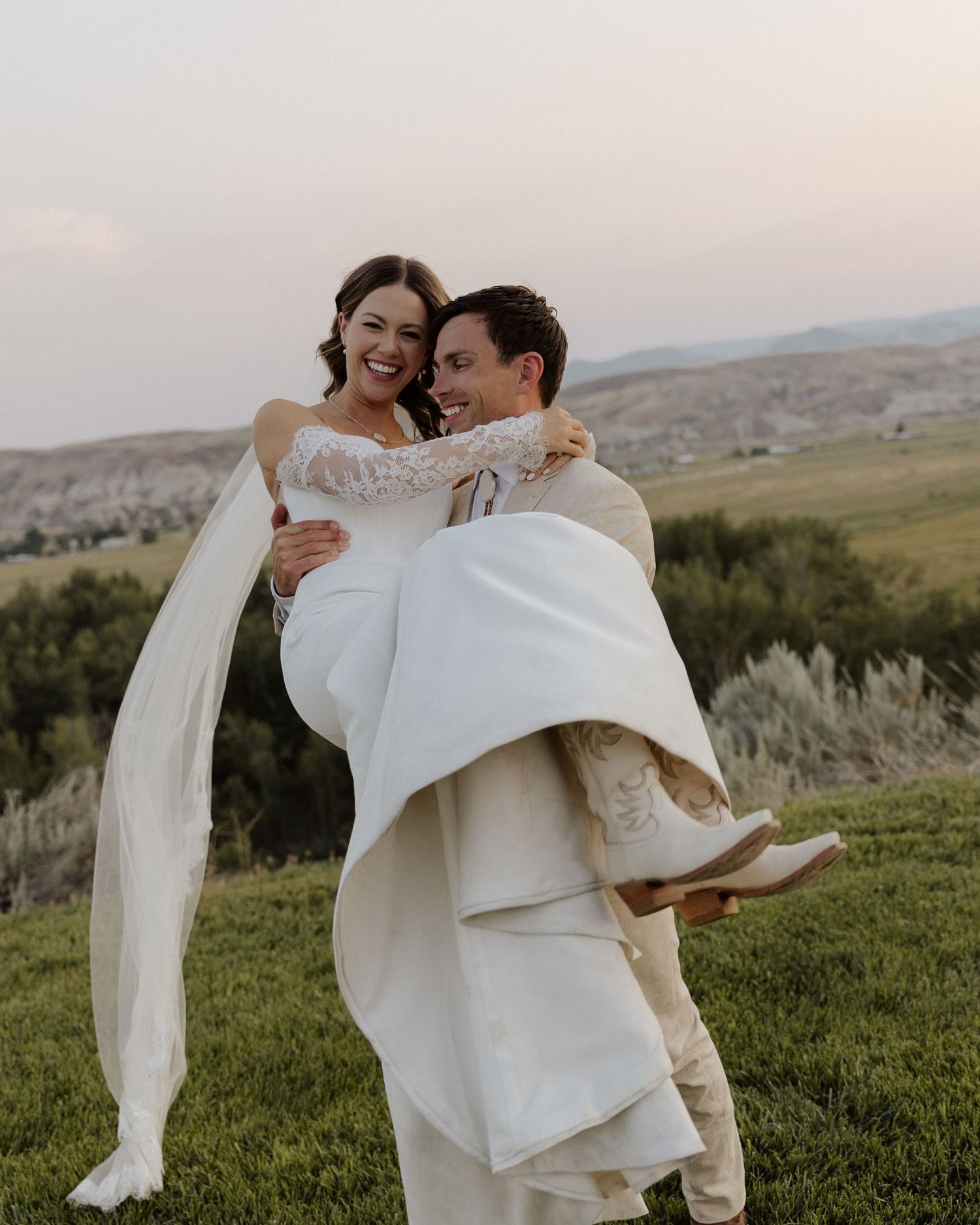 Groom carries bride in flowing white dress against sunset mountain backdrop.